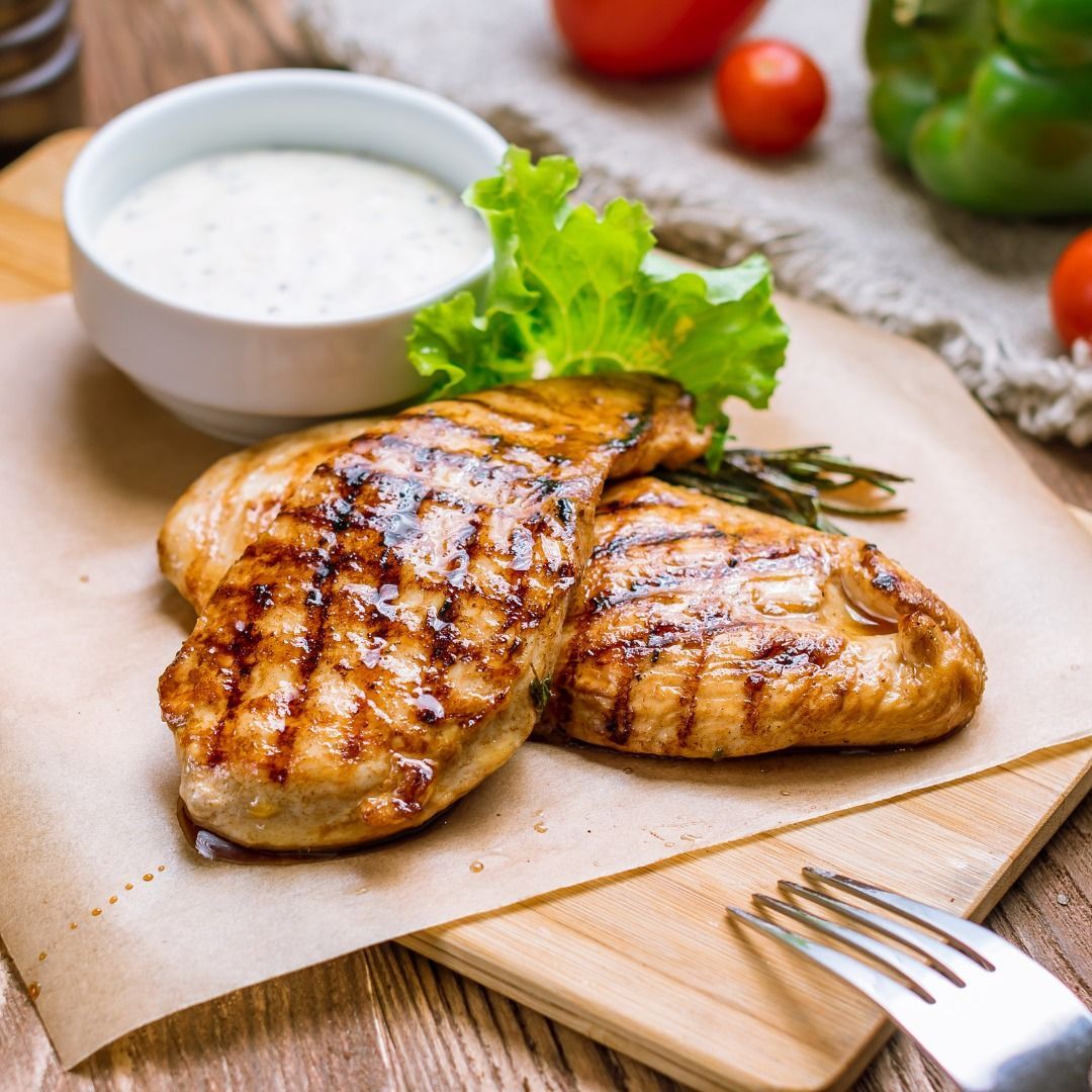 Grilled chicken breasts with lettuce and dipping sauce on a wooden board, with fork and vegetables nearby