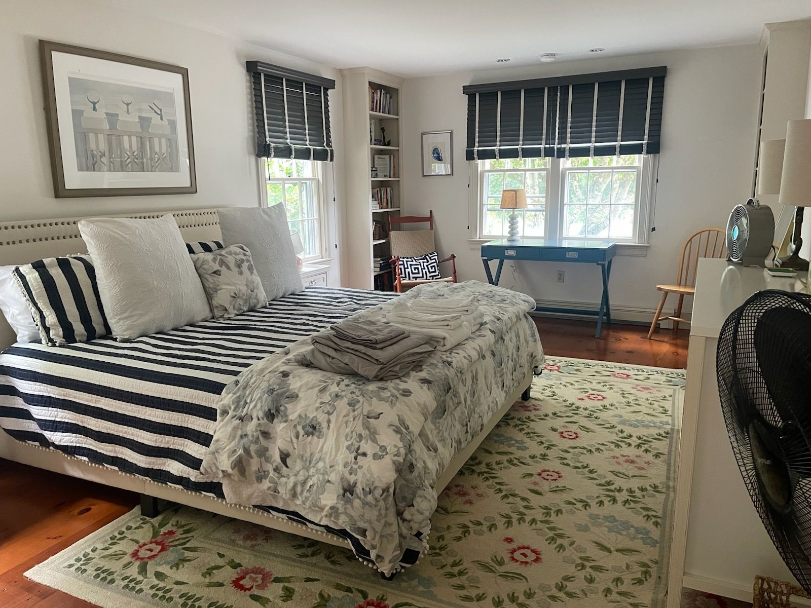 Bright bedroom with striped bed, floral rug, desk by windows, and a fan in the corner