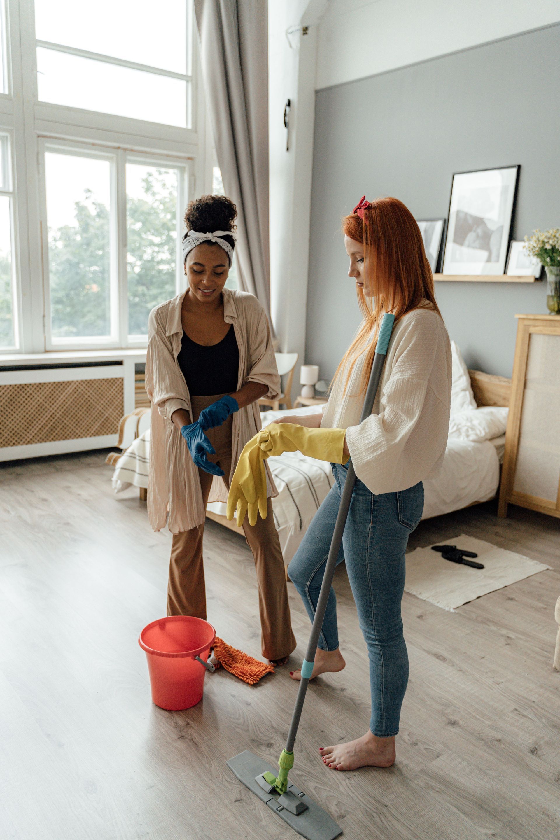 Two women mopping a bright living room, with a red bucket and yellow cloths on the floor.