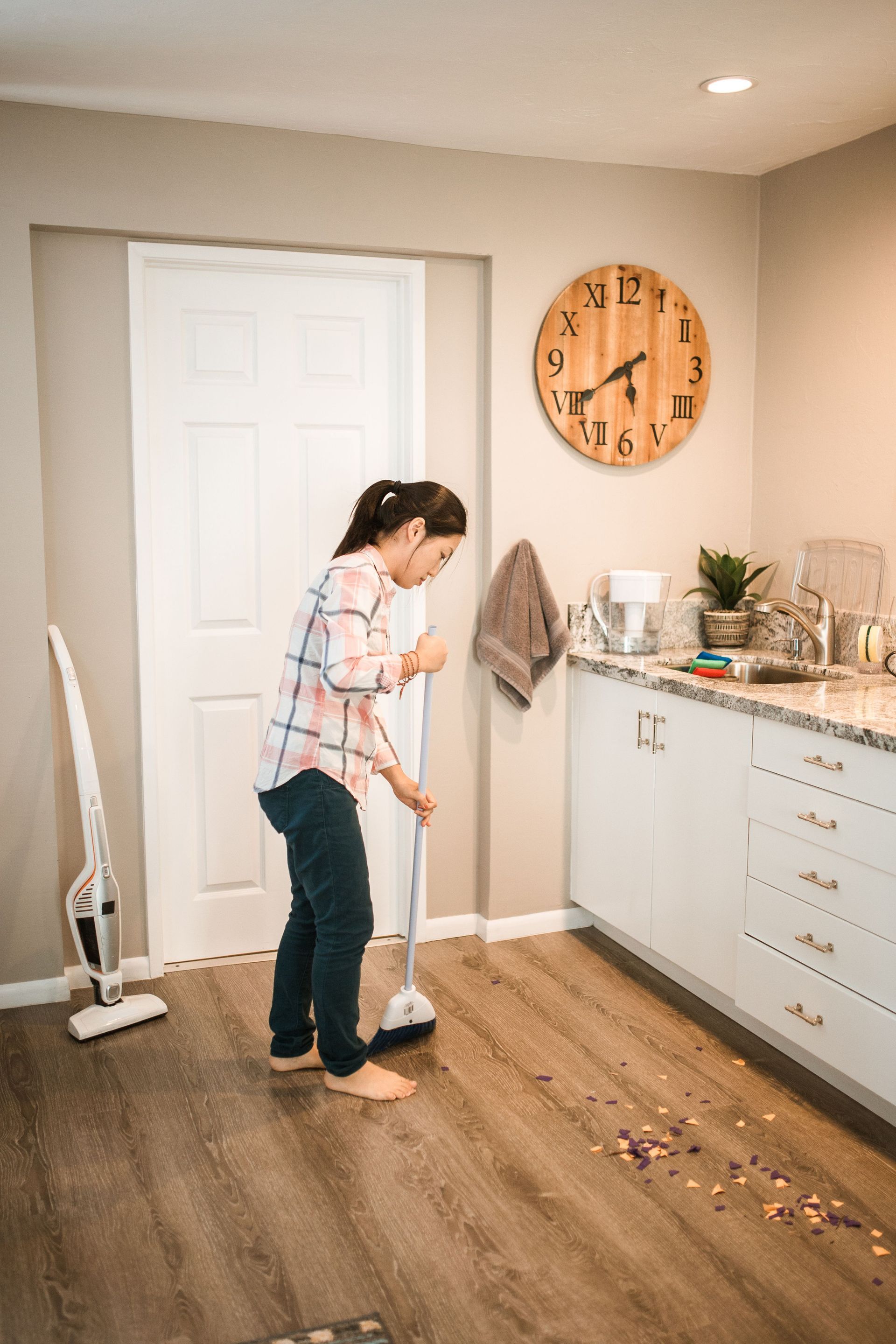 Person sweeping spilled cereal in a kitchen with white cabinets and a wooden wall clock.