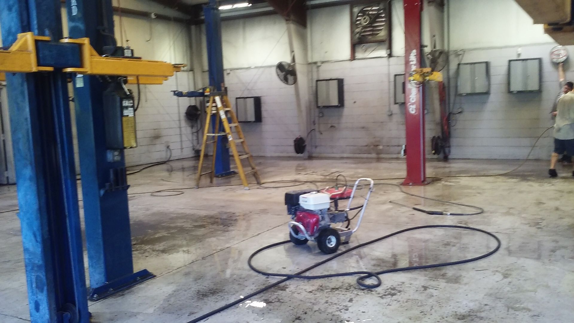 A power washer in a garage, cleaning the concrete floor. Blue lift, red pillar, and person visible. | Sloan's Automotive
