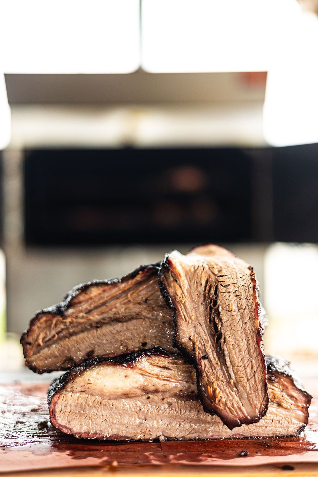 Stacked smoked meat slices on a wooden board, with a blurred kitchen or grill in the background