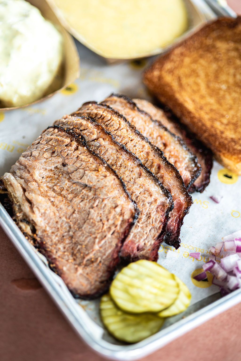 Sliced smoked brisket with pickles and cornbread on a tray, served with sides.