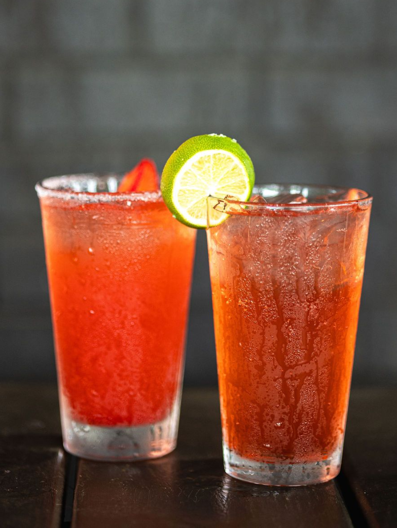 Two red cocktails in tall glasses, one garnished with a lime slice on a dark table