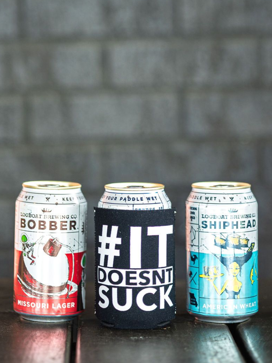 Three beverage cans on a surface, including one with “#IT DOESN’T SUCK,” against a gray wall.