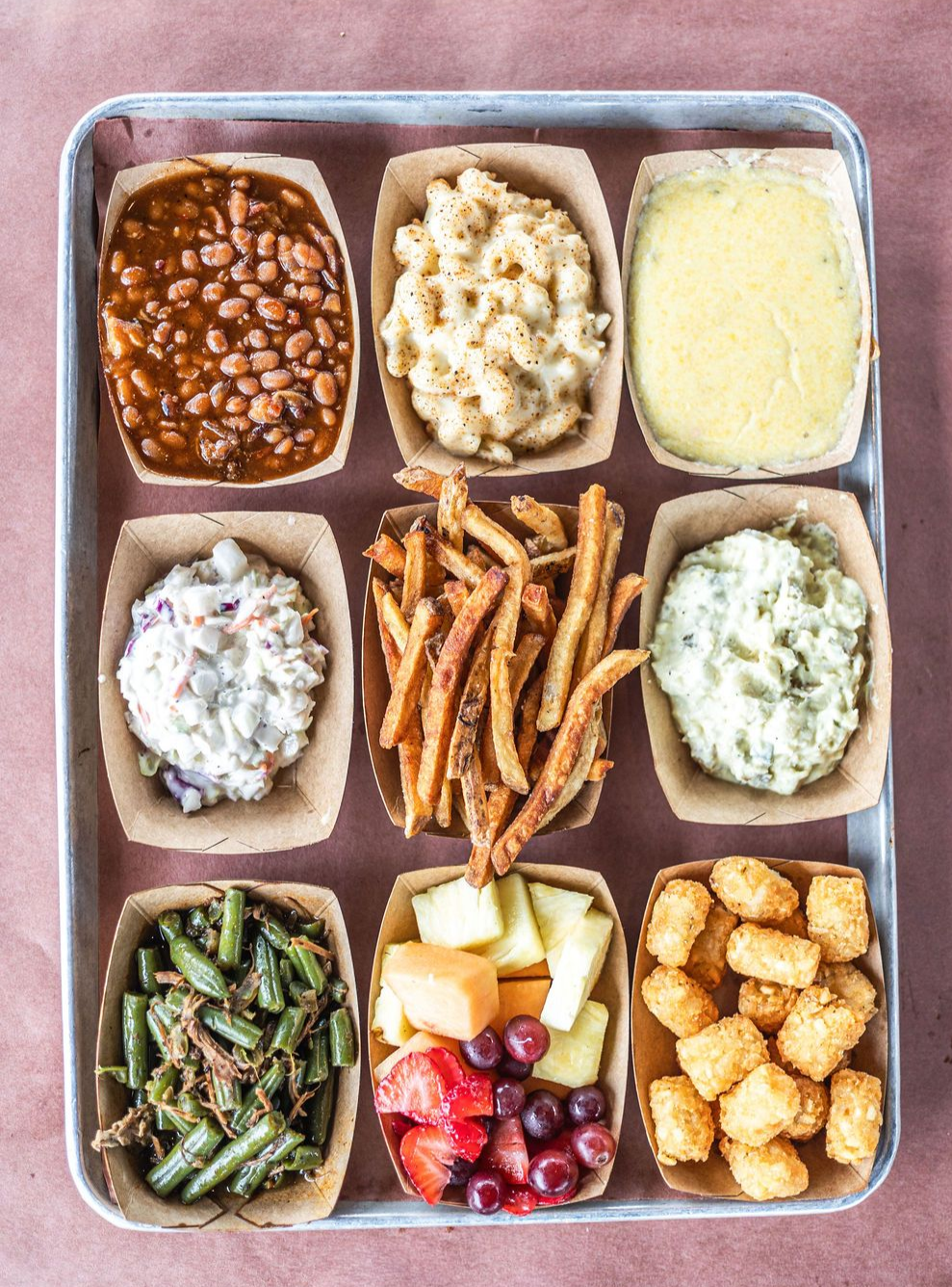 Assorted takeout meal tray with nine compartments of beans, mashed potatoes, fries, salad, green beans, fruit, and tater tots