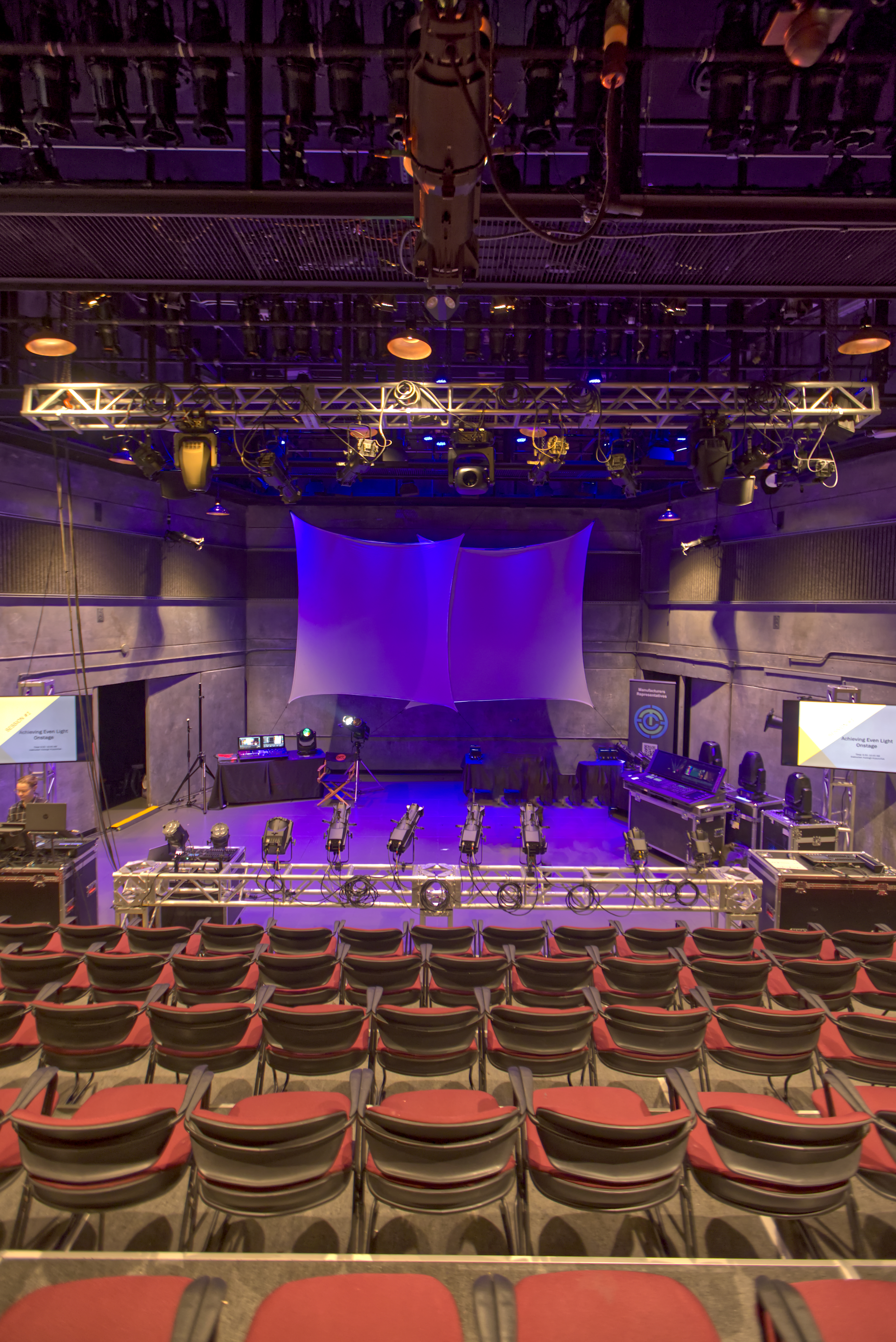 Empty theater auditorium with red chairs facing a lit stage and lighting fixtures.