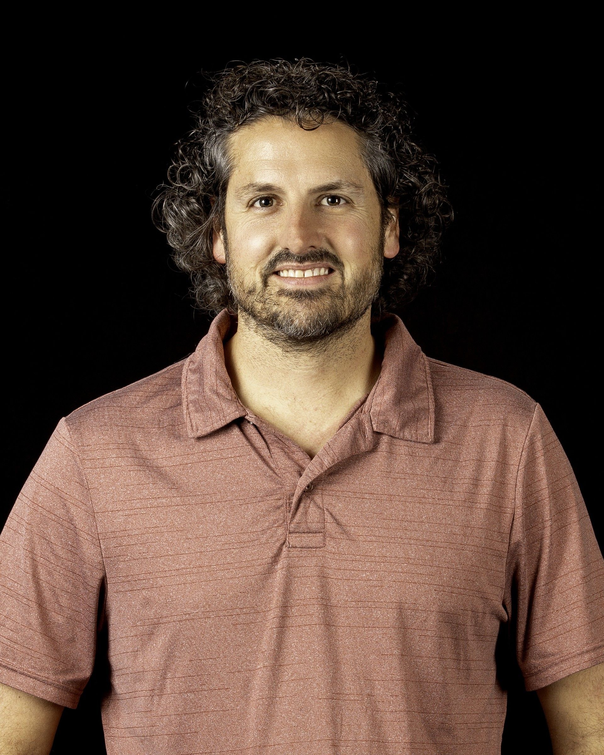 Man with curly brown hair and a beard smiles, wearing a red polo shirt, against a black background.