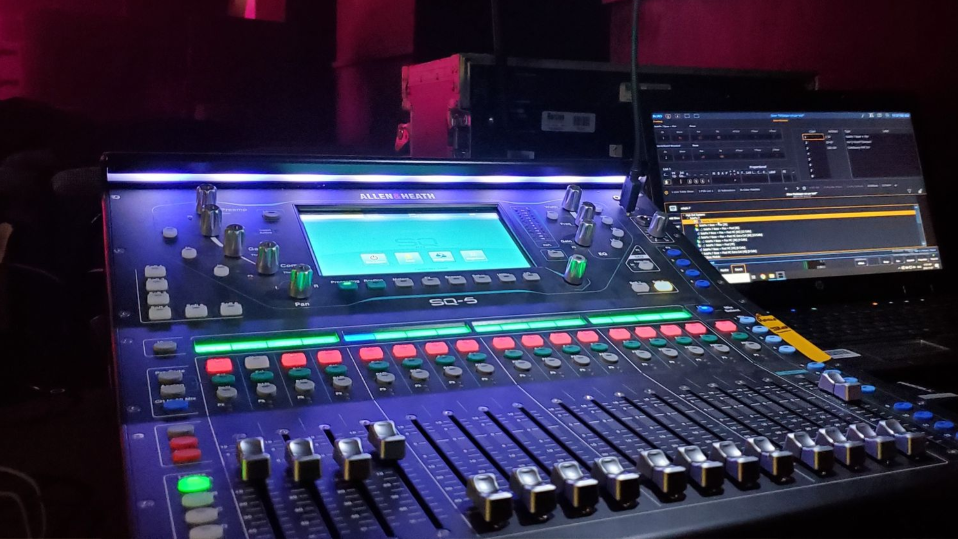 Sound mixing console with illuminated controls and a connected tablet, likely in a dark venue.