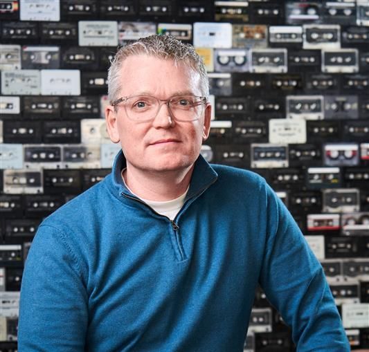 Man with glasses in blue sweater, smiling, against a backdrop of audio cassettes.