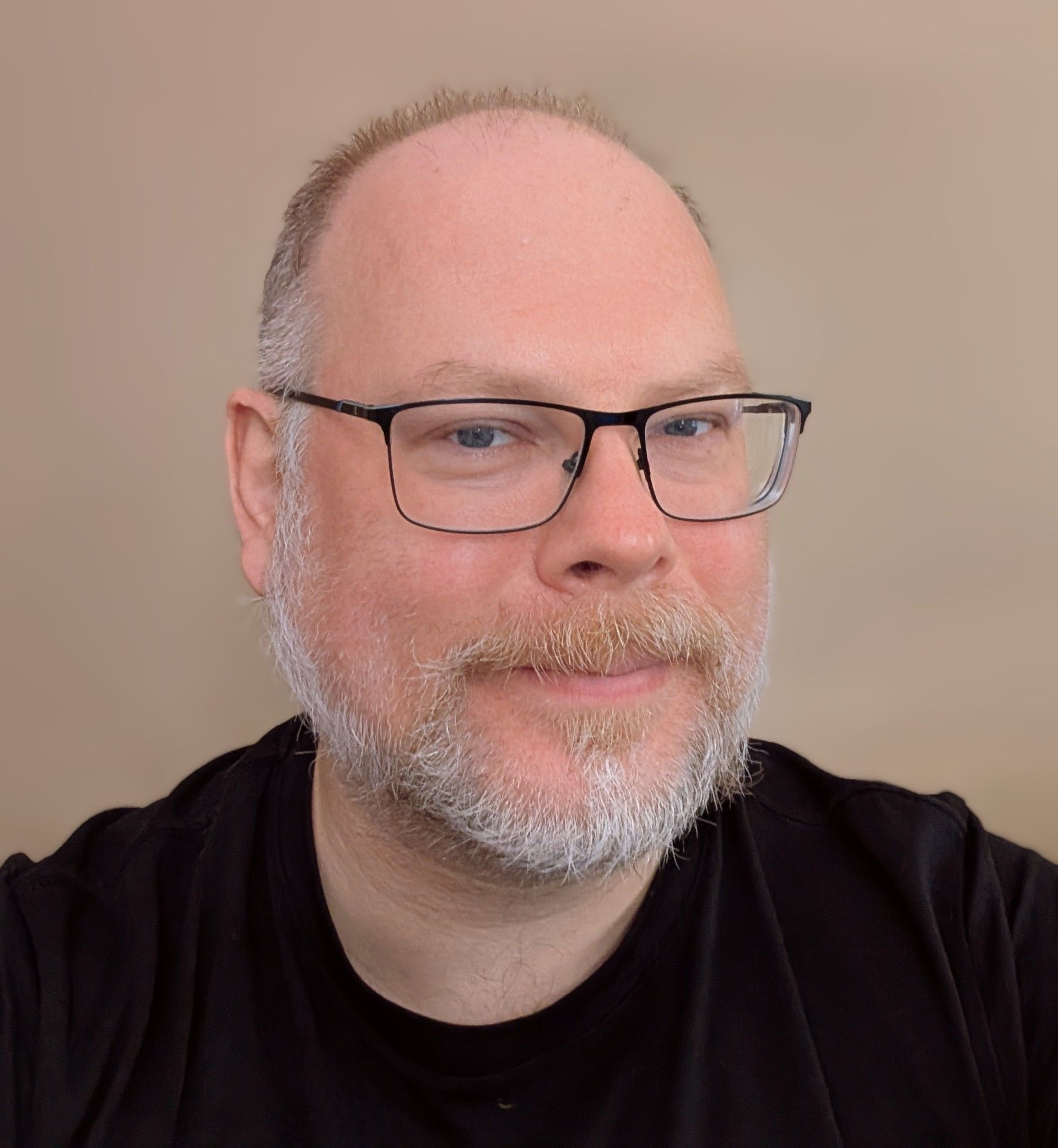 Man with glasses and a beard smiling, wearing a black shirt, in front of a neutral background.