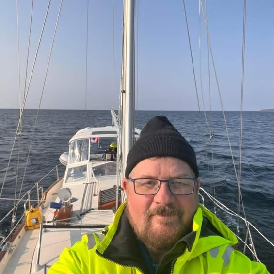 Man on sailboat, wearing a black hat and yellow jacket, sailing on calm water under a blue sky.