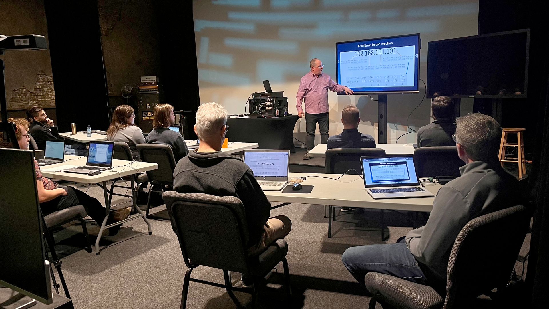 Man presenting to a group in a dimly lit room with screens and laptops. The presenter points to a screen.