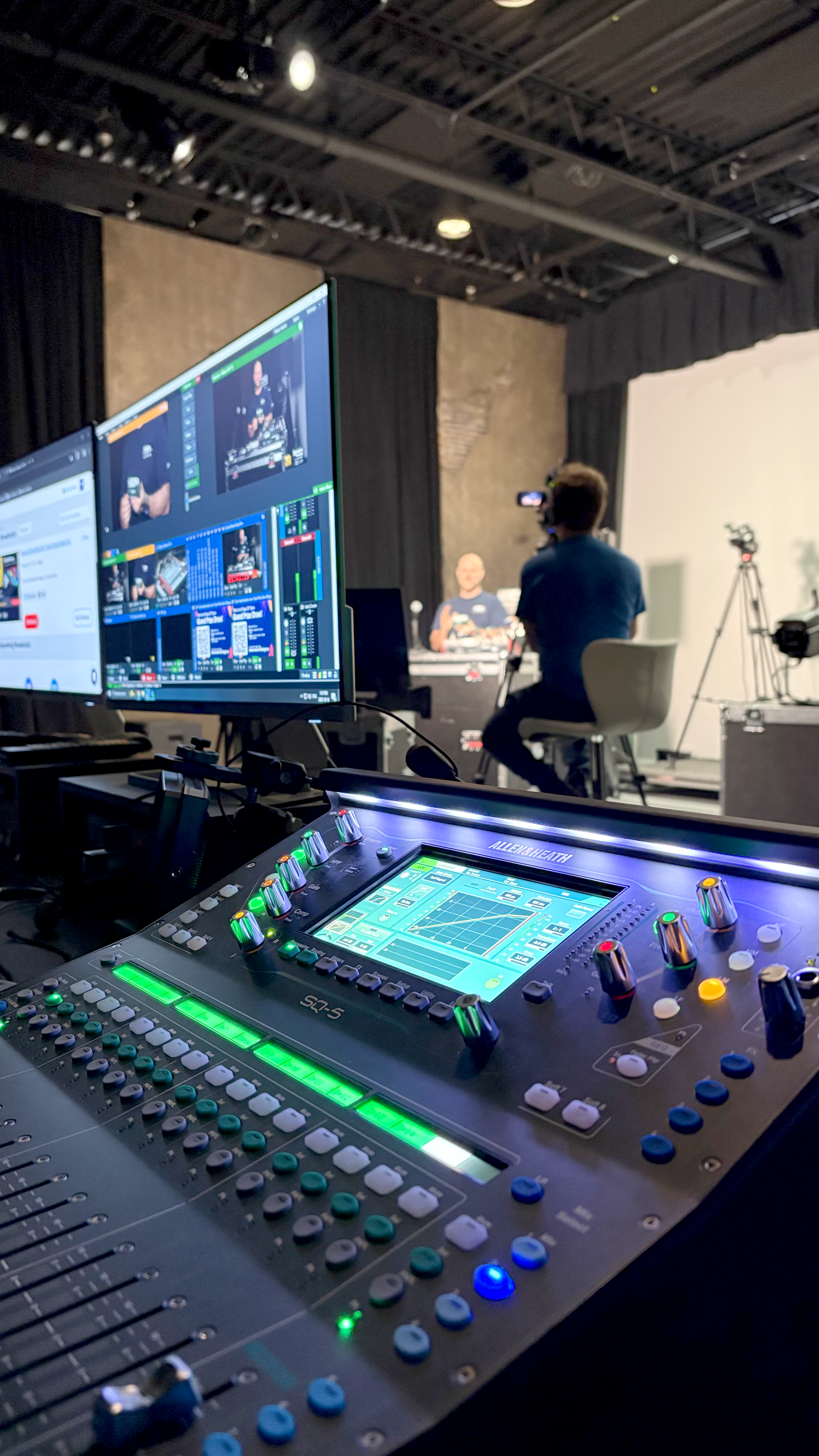 Audio mixing console with screens displaying a live video production. A person films the interview set.