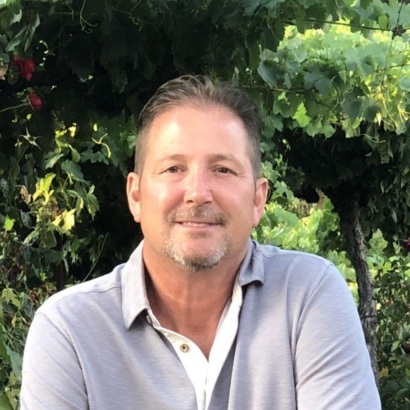 Man in gray polo shirt smiles in front of green foliage.