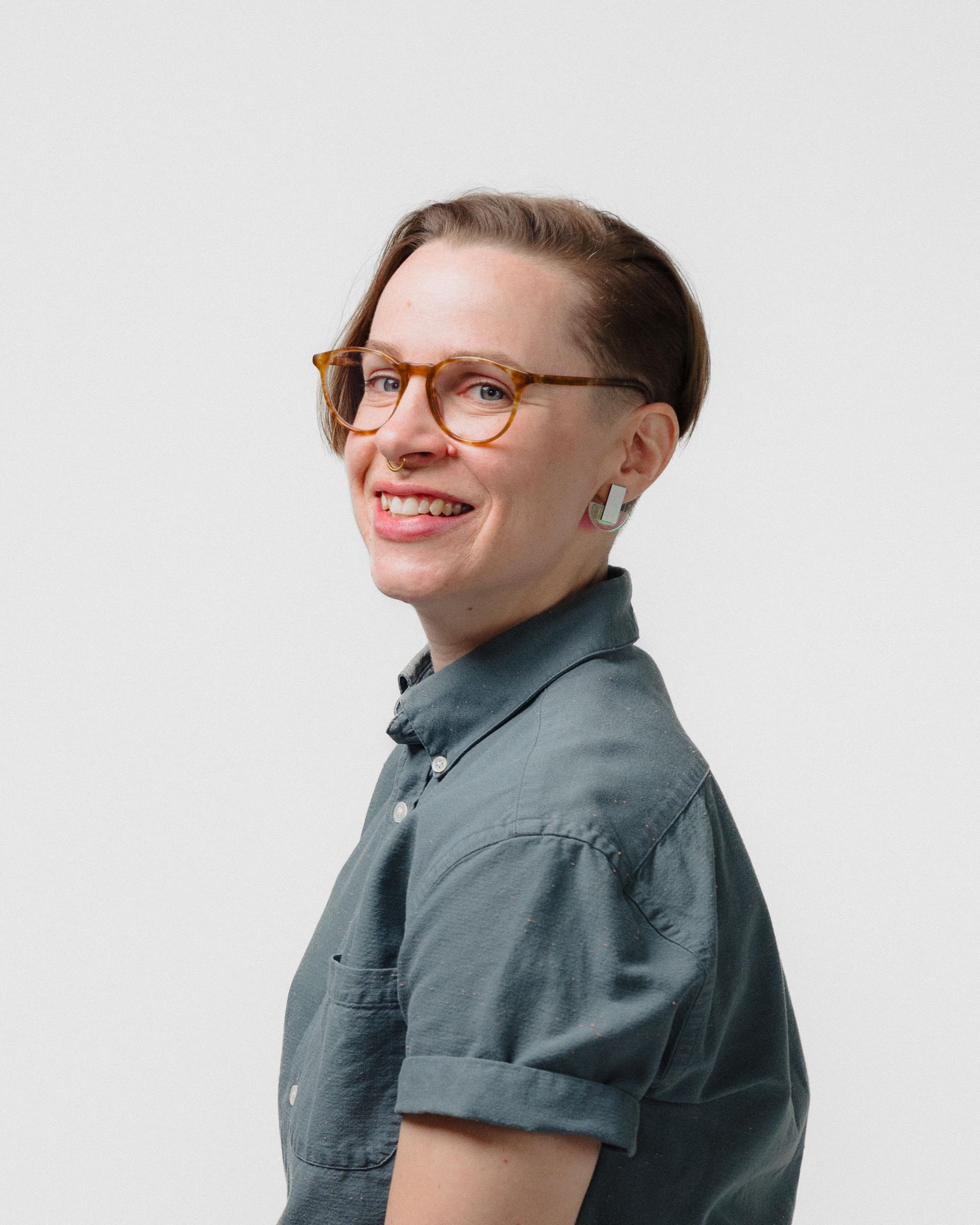 Woman with glasses smiling, wearing a blue shirt, looking over her shoulder.