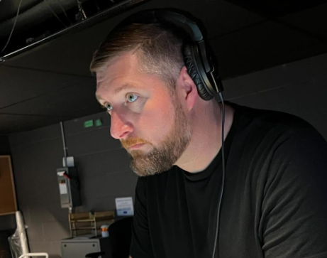 Man with headphones, looking intently. Dark setting, wearing a black shirt.