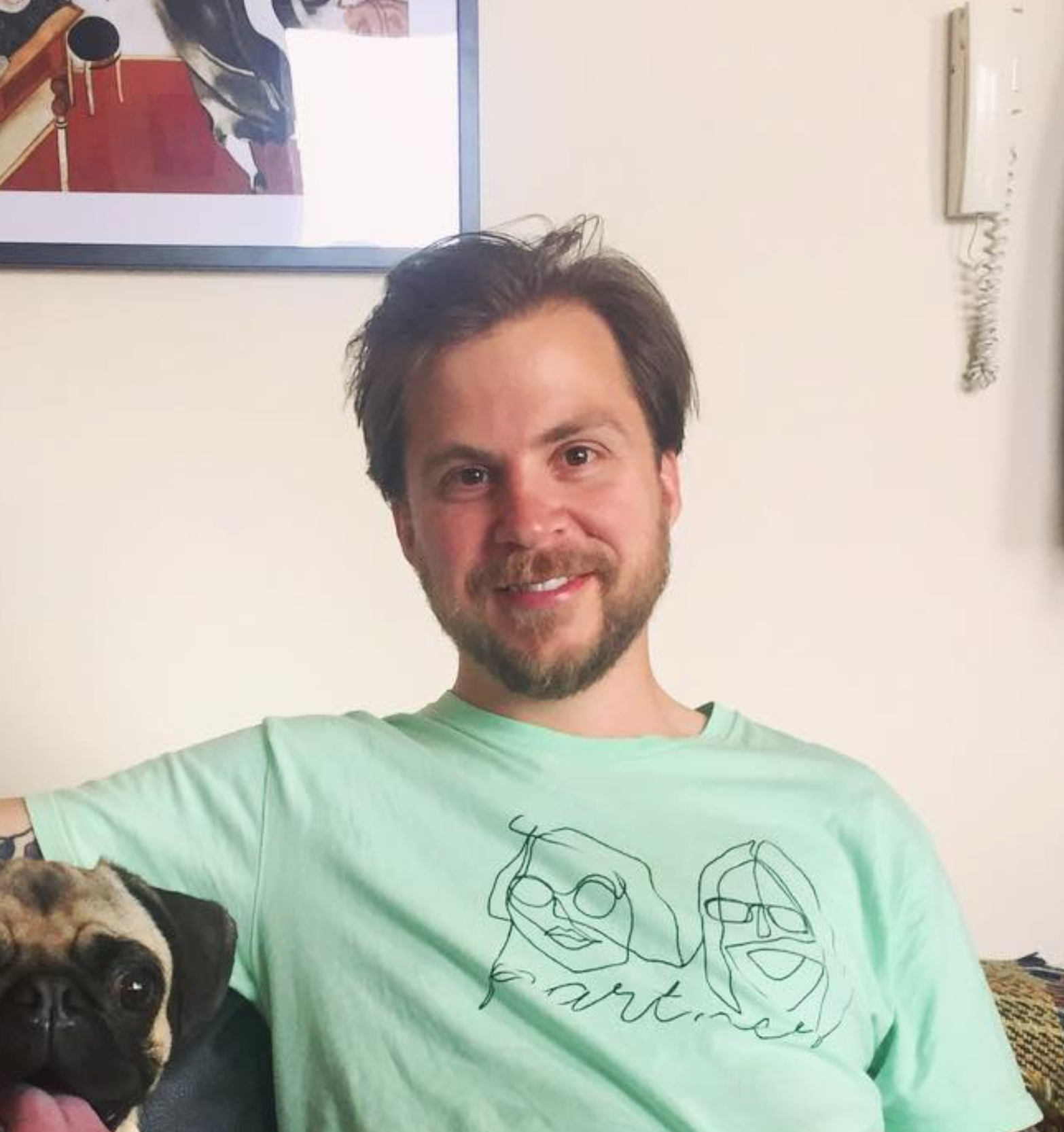 Man in light green shirt smiles next to a pug. A framed picture and a phone are on the wall.