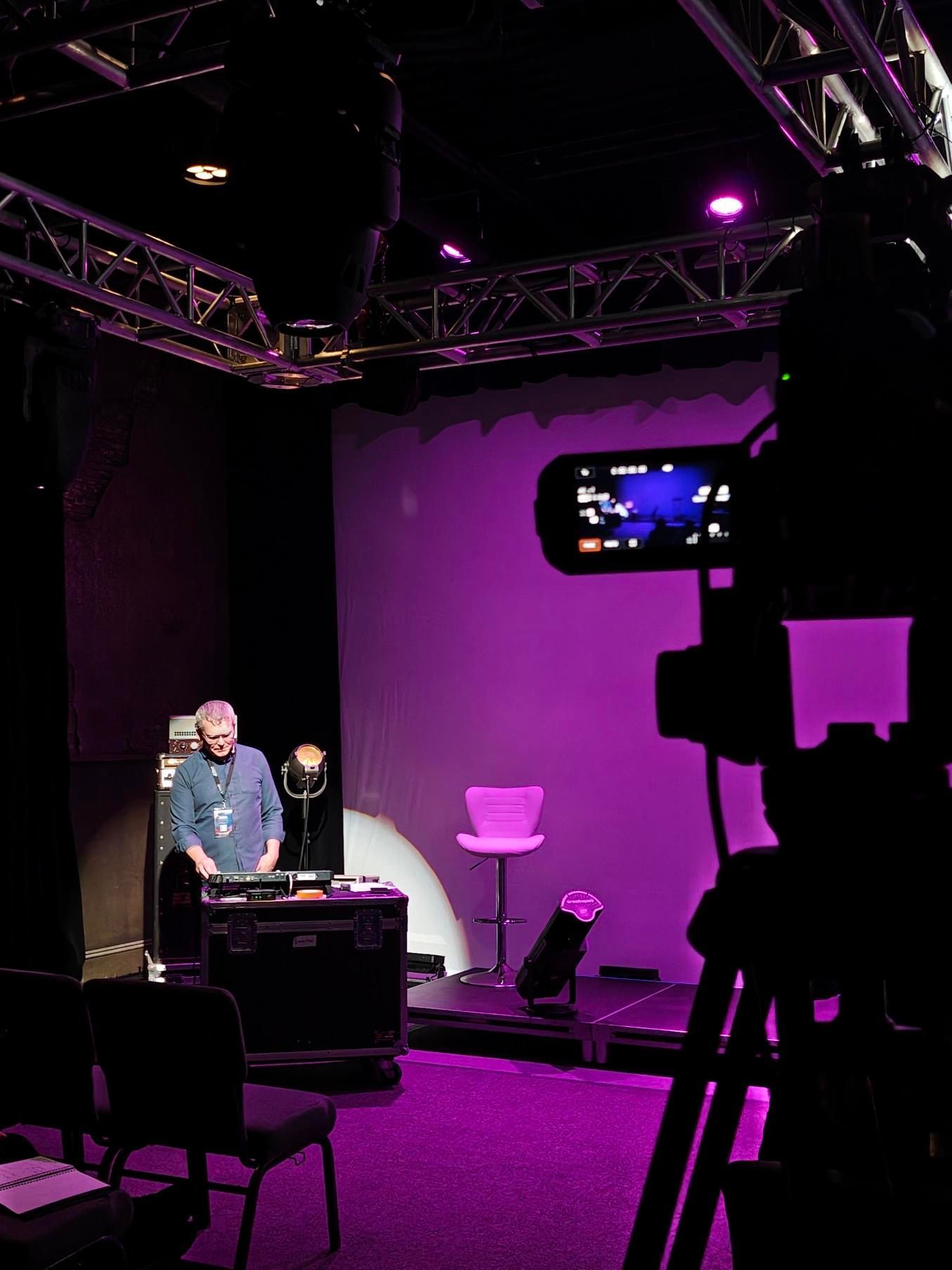 Man at a music setup in a studio, illuminated by purple light, with a camera capturing the scene.