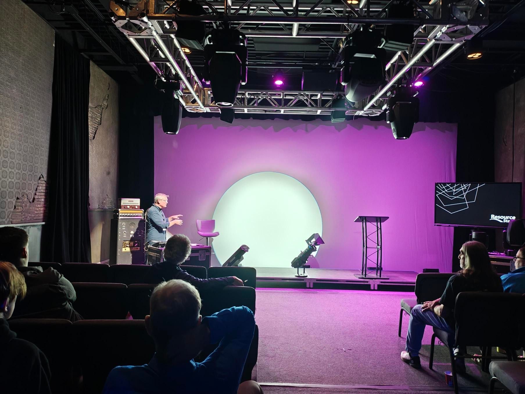 A man speaks on a stage with a purple backdrop and audience. Stage lights shine on a white circle.