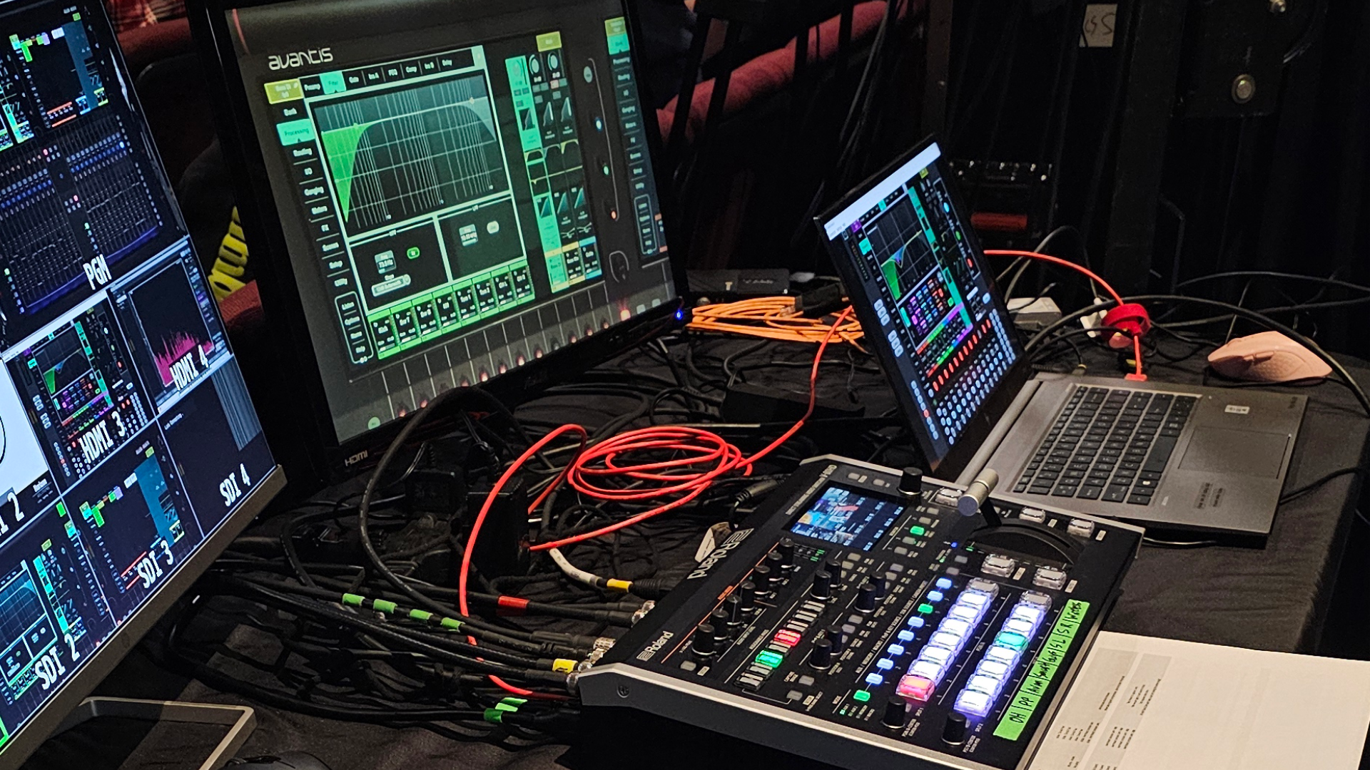 Sound mixing console and screens displaying audio levels, connected by cables, in a dark room.