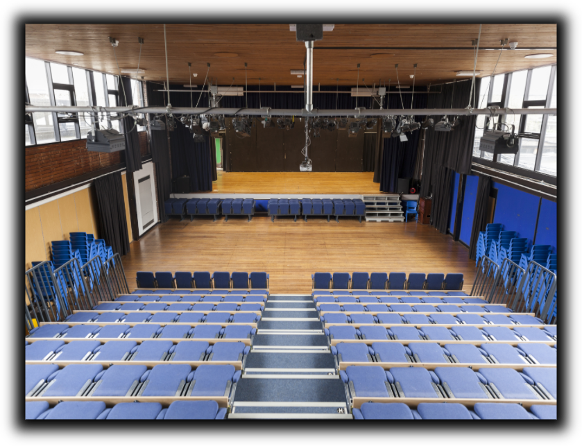 Auditorium with blue seating facing a wooden stage. Overhead lights and stage curtains are visible.