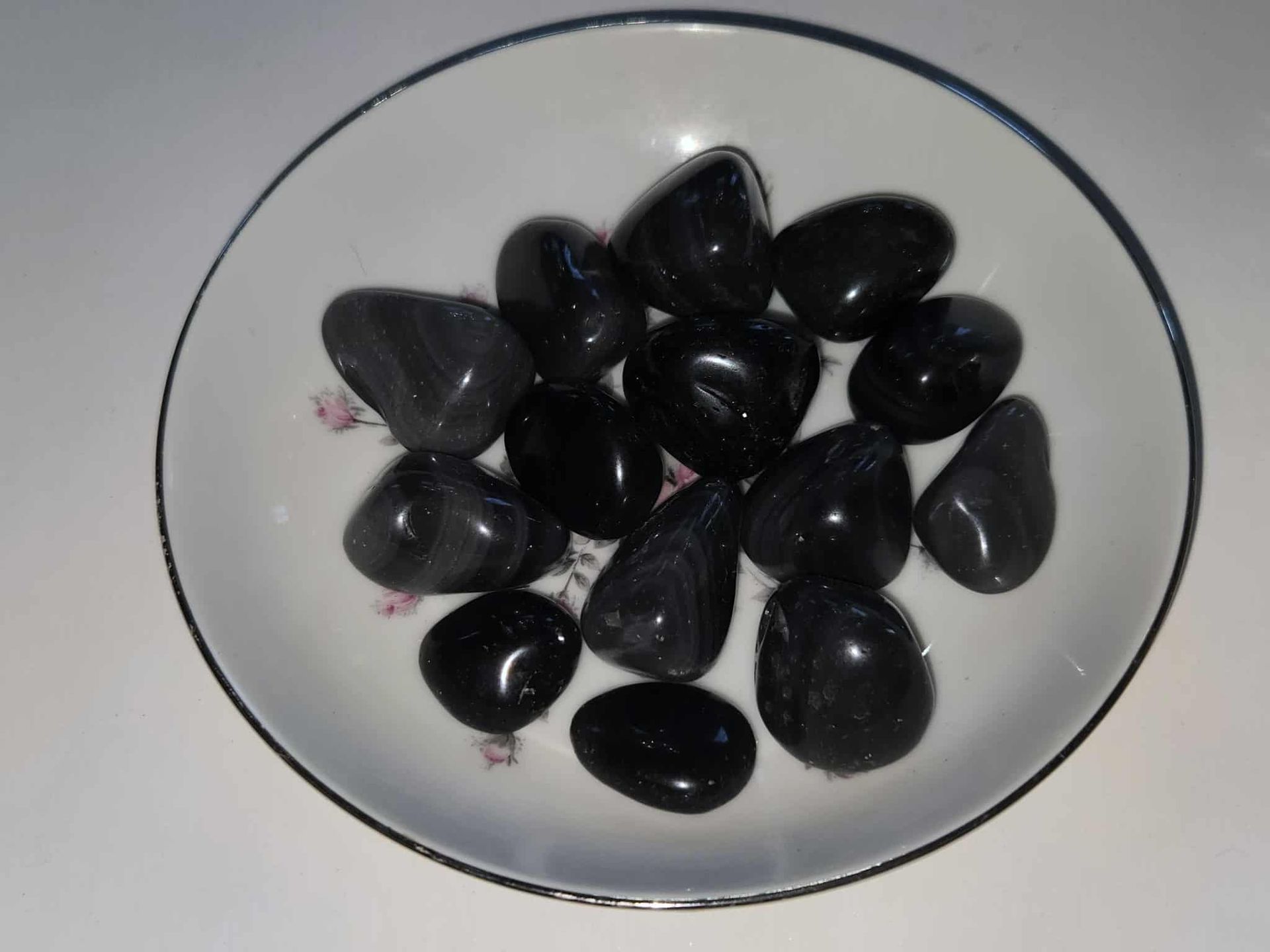 Black, polished stones in a white floral-decorated bowl, set on a white surface.