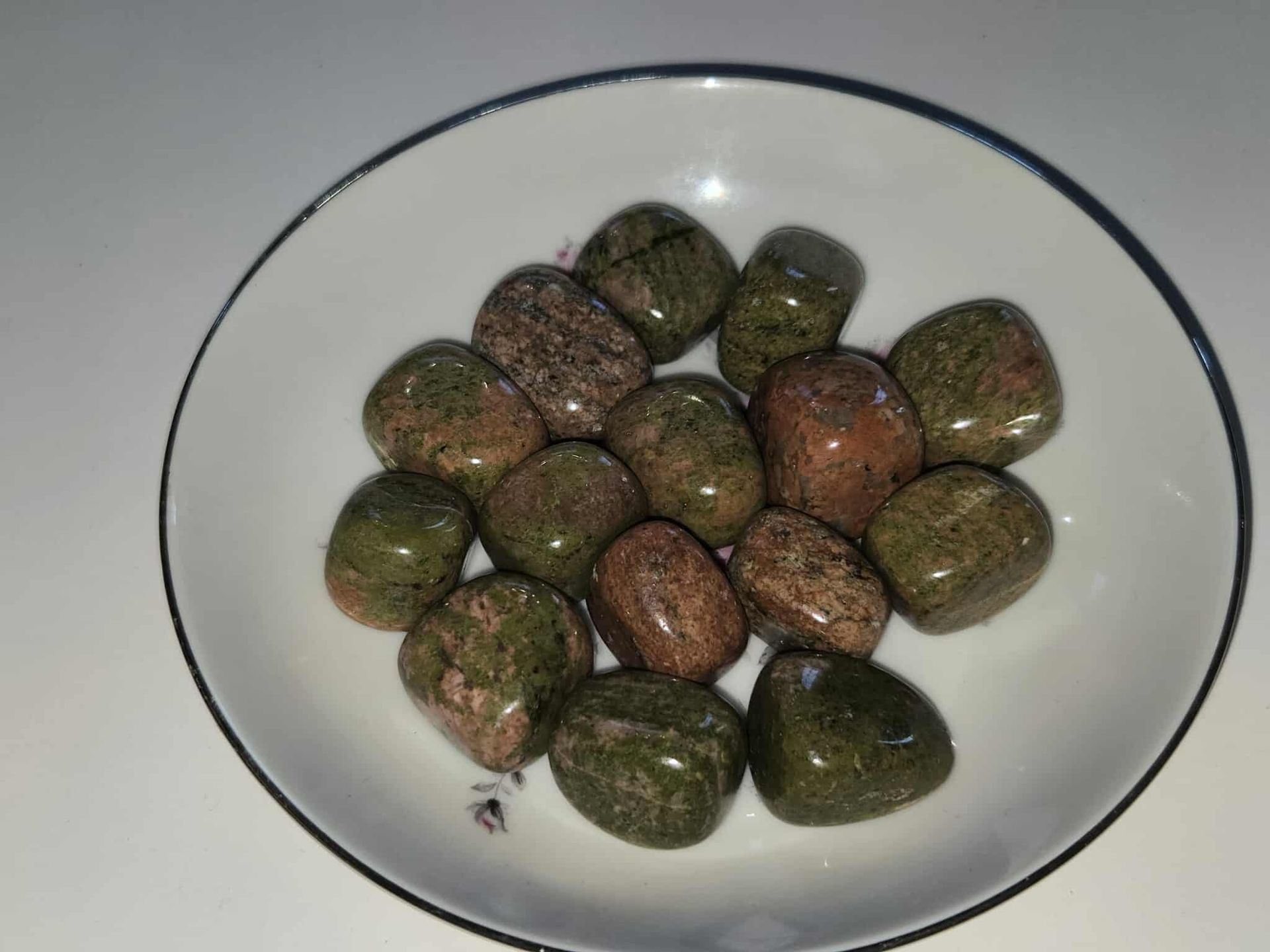Tumbled unakite stones, green and pink hues, arranged in a white bowl with a blue rim.