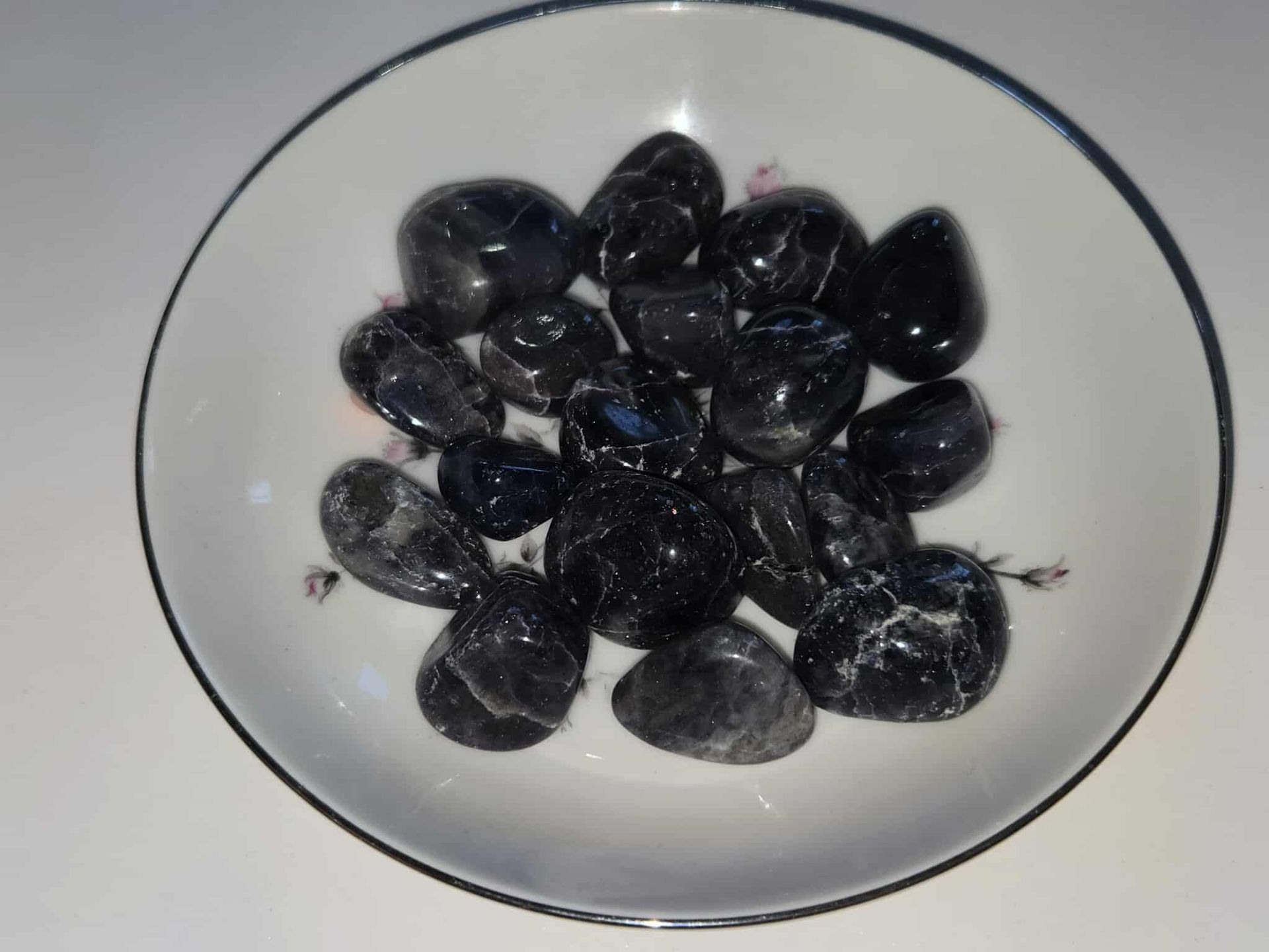 Black tumbled stones in a white floral-decorated bowl on a white surface.