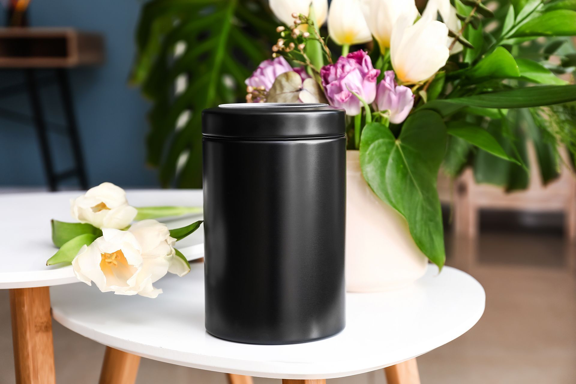 Black urn on a white table with flowers; room interior with a blurred background.