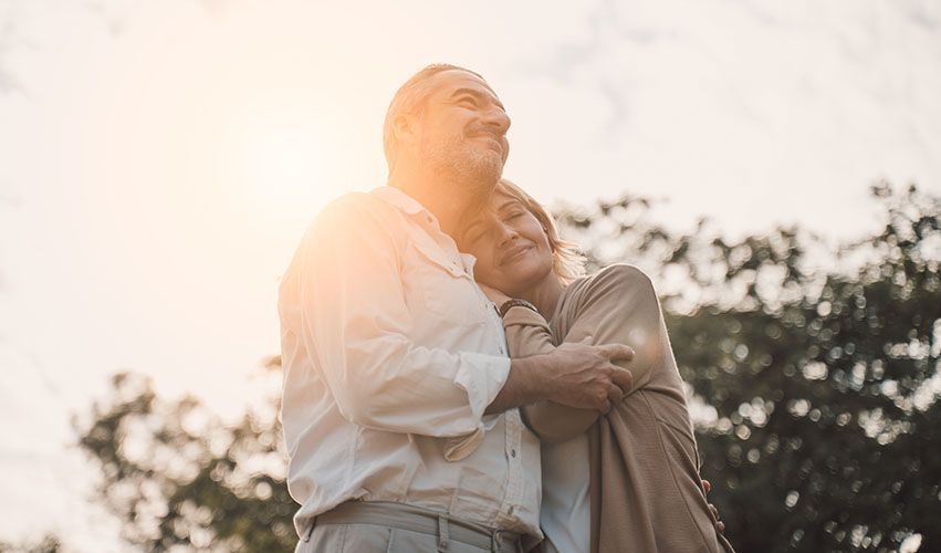 Older couple embracing outdoors in sunlight; woman resting head on man's shoulder.