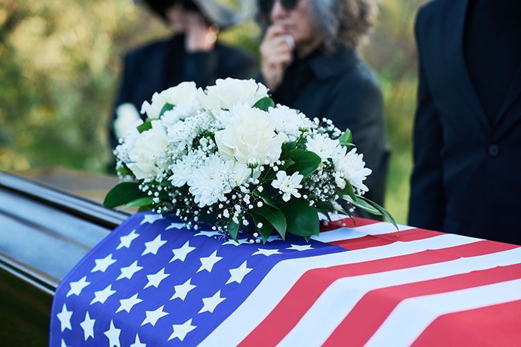Coffin draped with American flag, topped with white flower arrangement; people at a funeral.