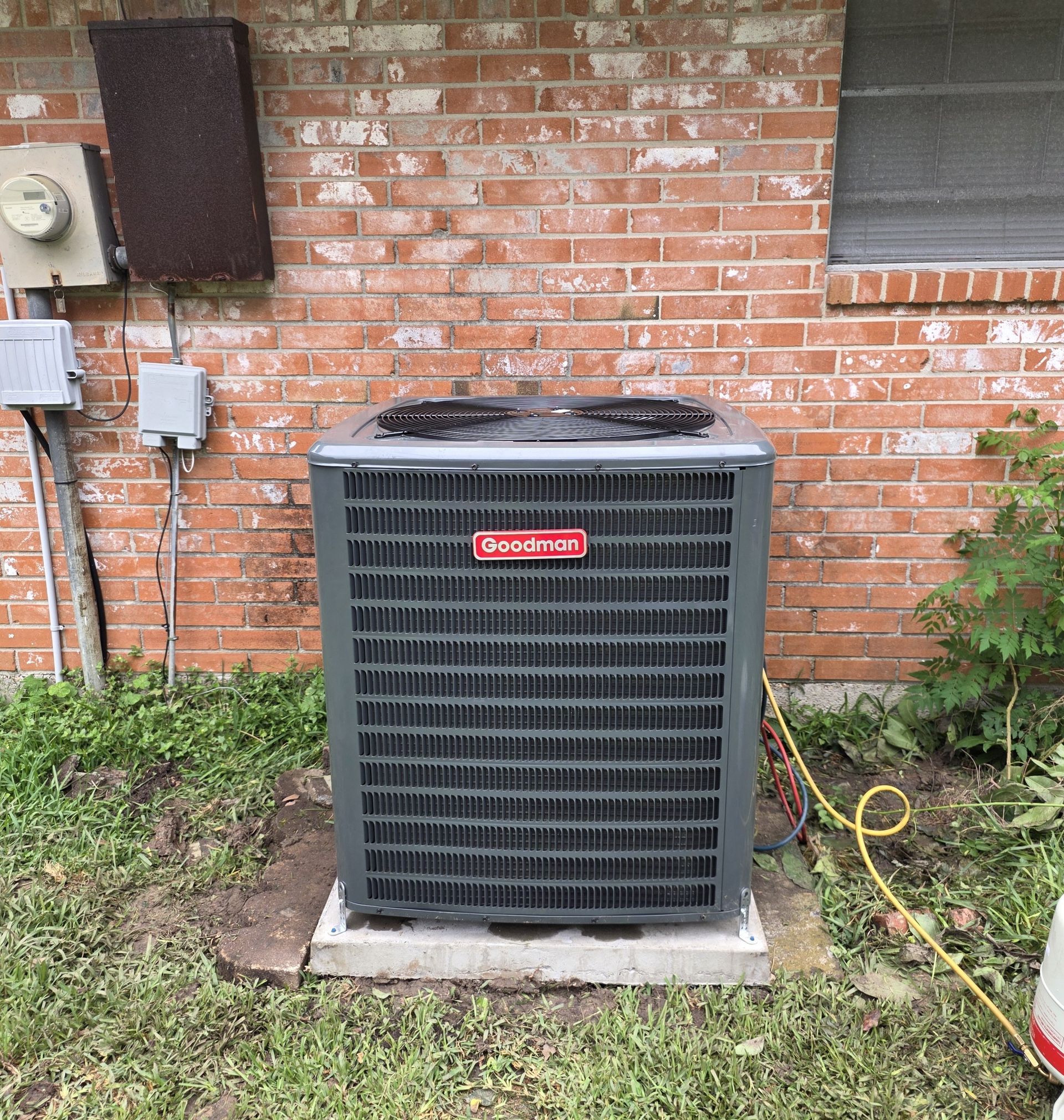 Outdoor Goodman air conditioner unit beside a brick house, with grass and utility boxes nearby.