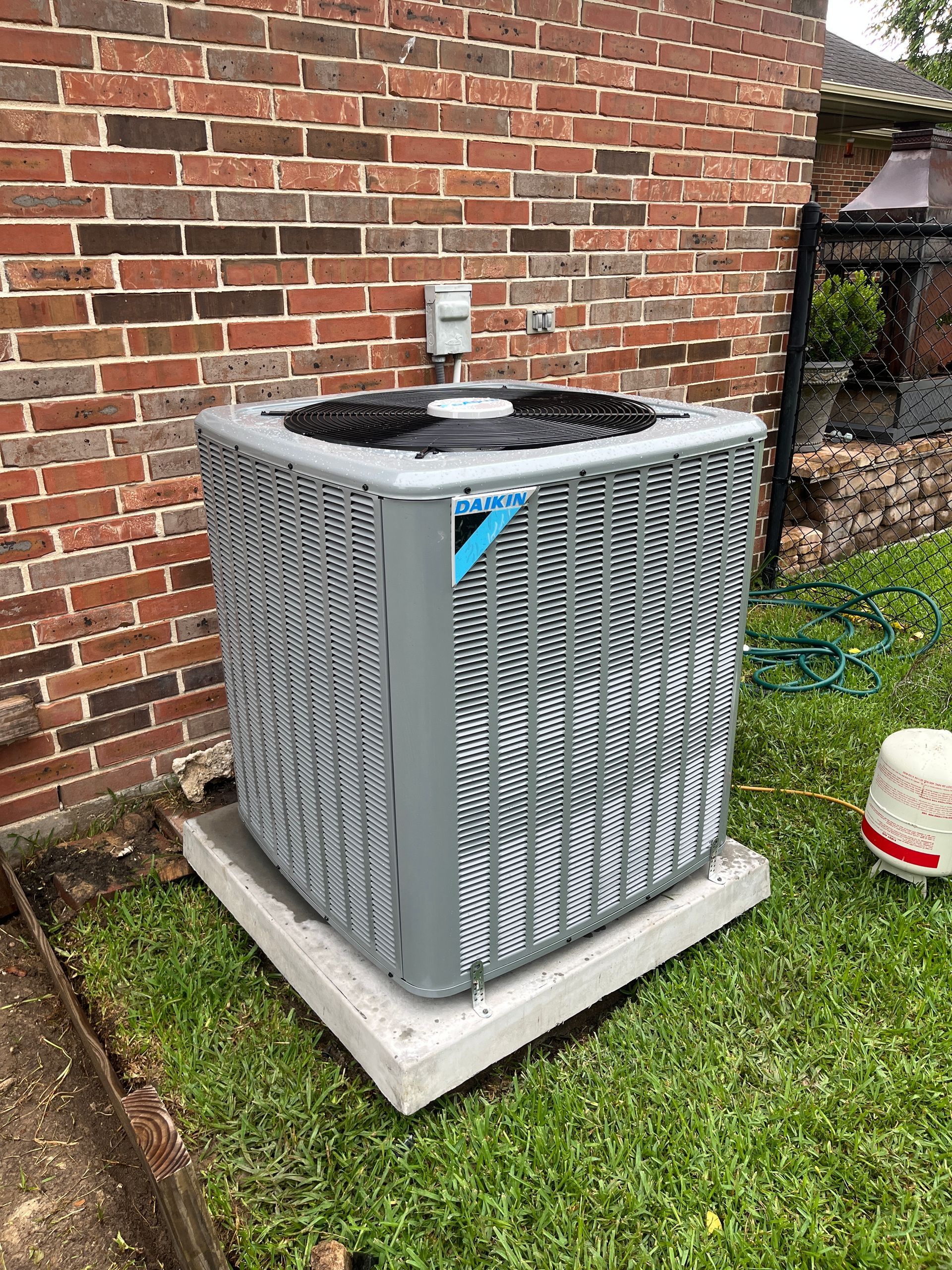 Outdoor HVAC unit beside a brick wall on a concrete pad, with grass and a utility marker nearby