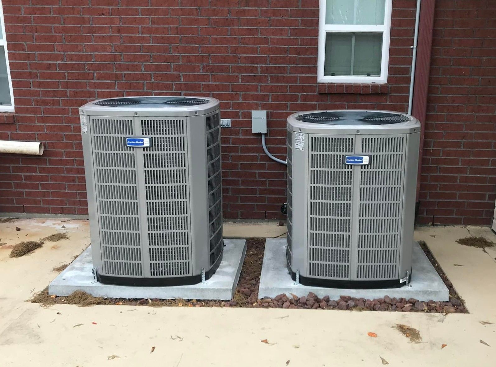 Two outdoor air conditioning units beside a brick wall on concrete pads