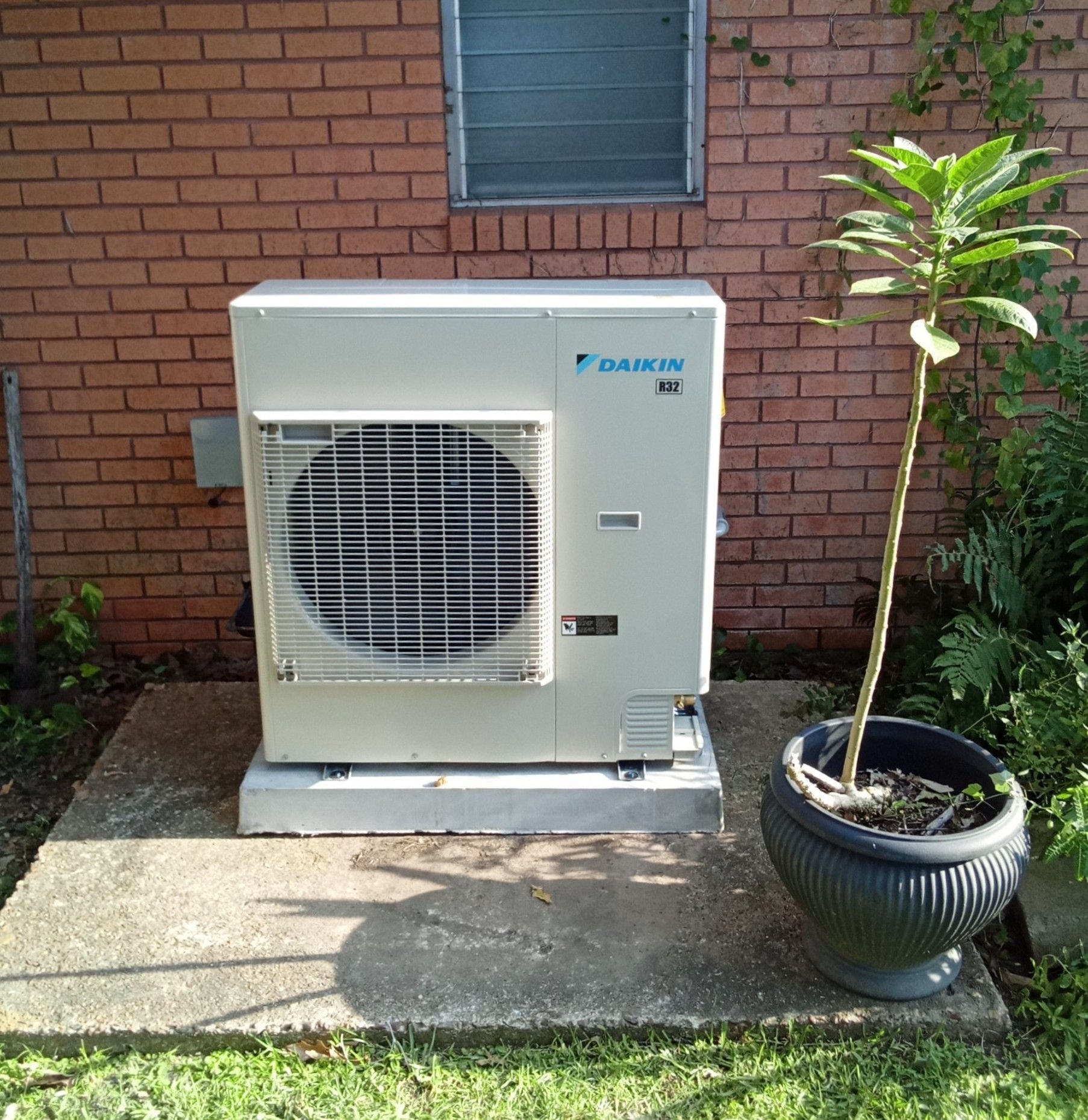 Outdoor air conditioner unit beside a brick wall, with a potted tree on a concrete patio.
