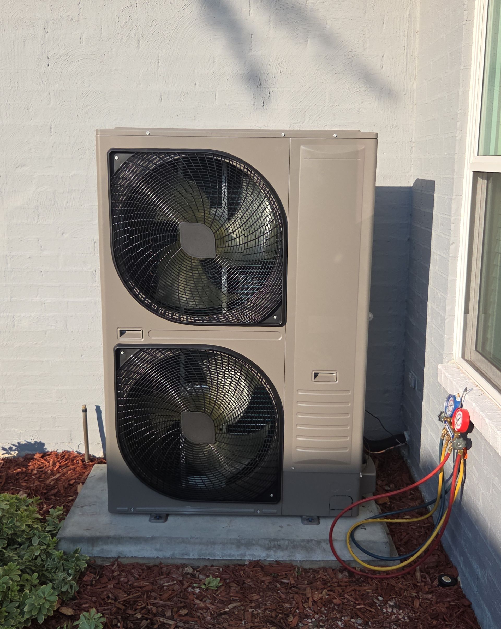 Outdoor heat pump unit beside a house wall, with hoses attached and sunlight casting shadows.