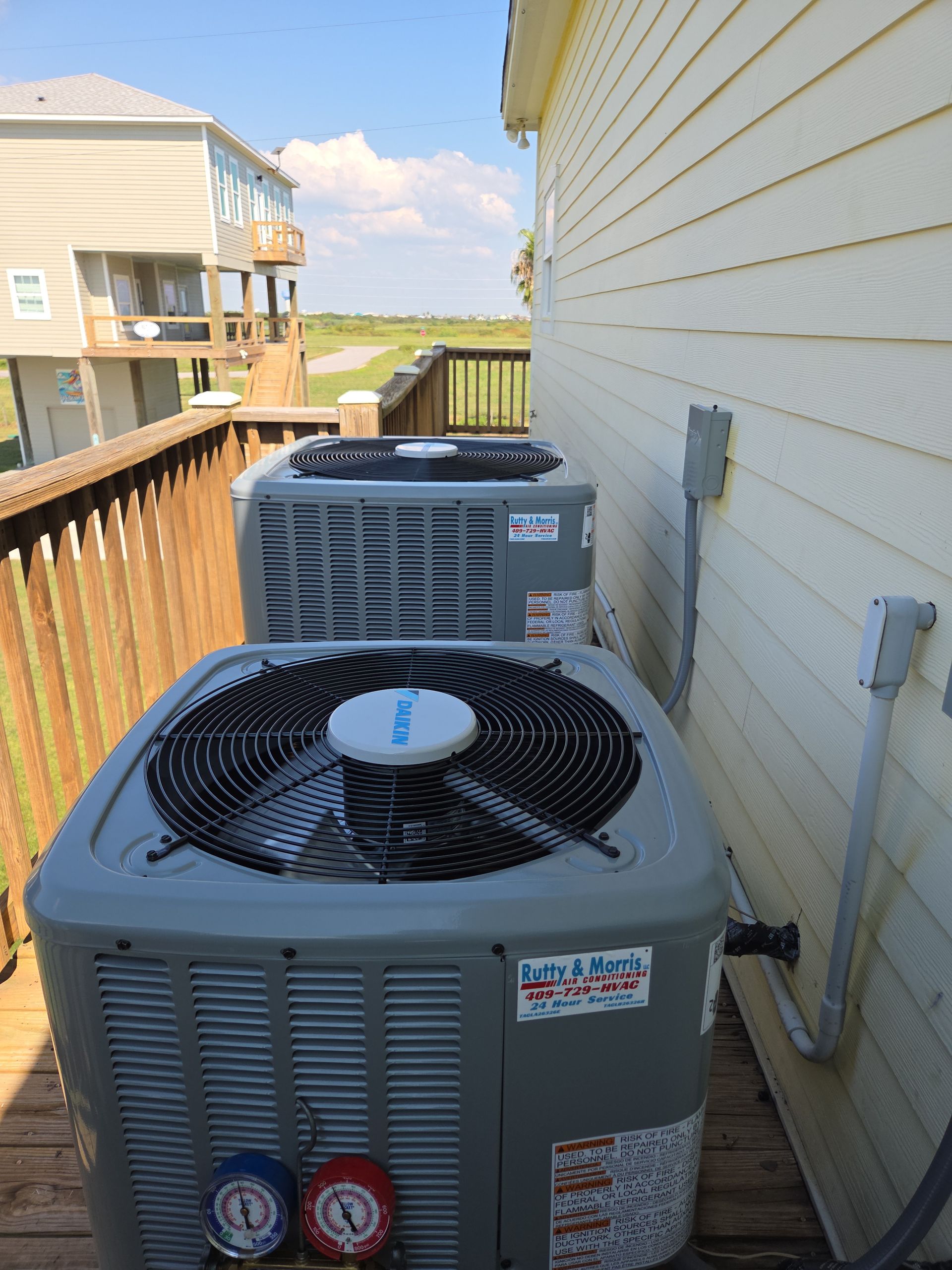 Two outdoor air conditioning units beside a house along a wooden deck walkway