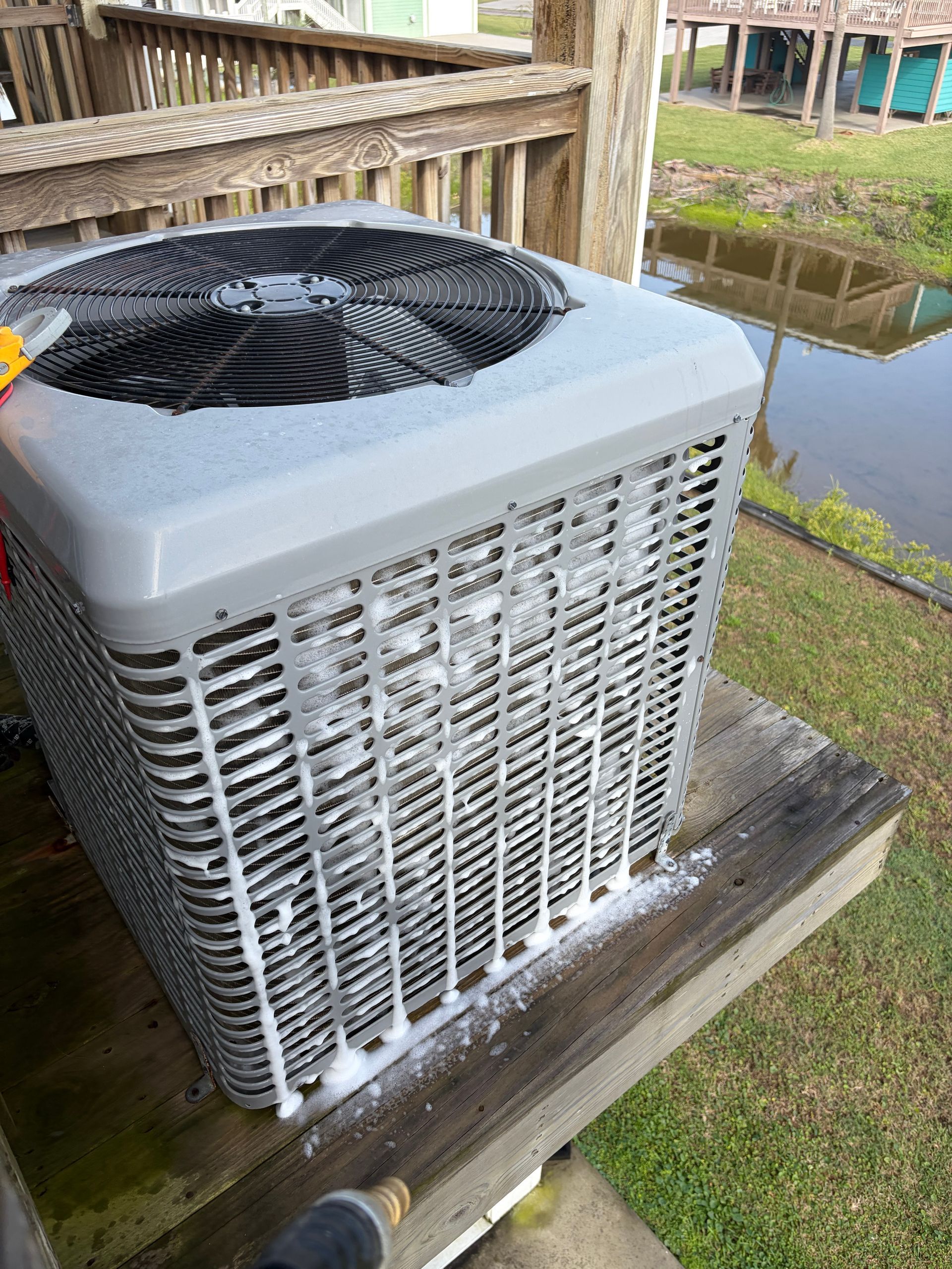 Outdoor HVAC unit beside a wooden deck, with a pond and fence in the background.