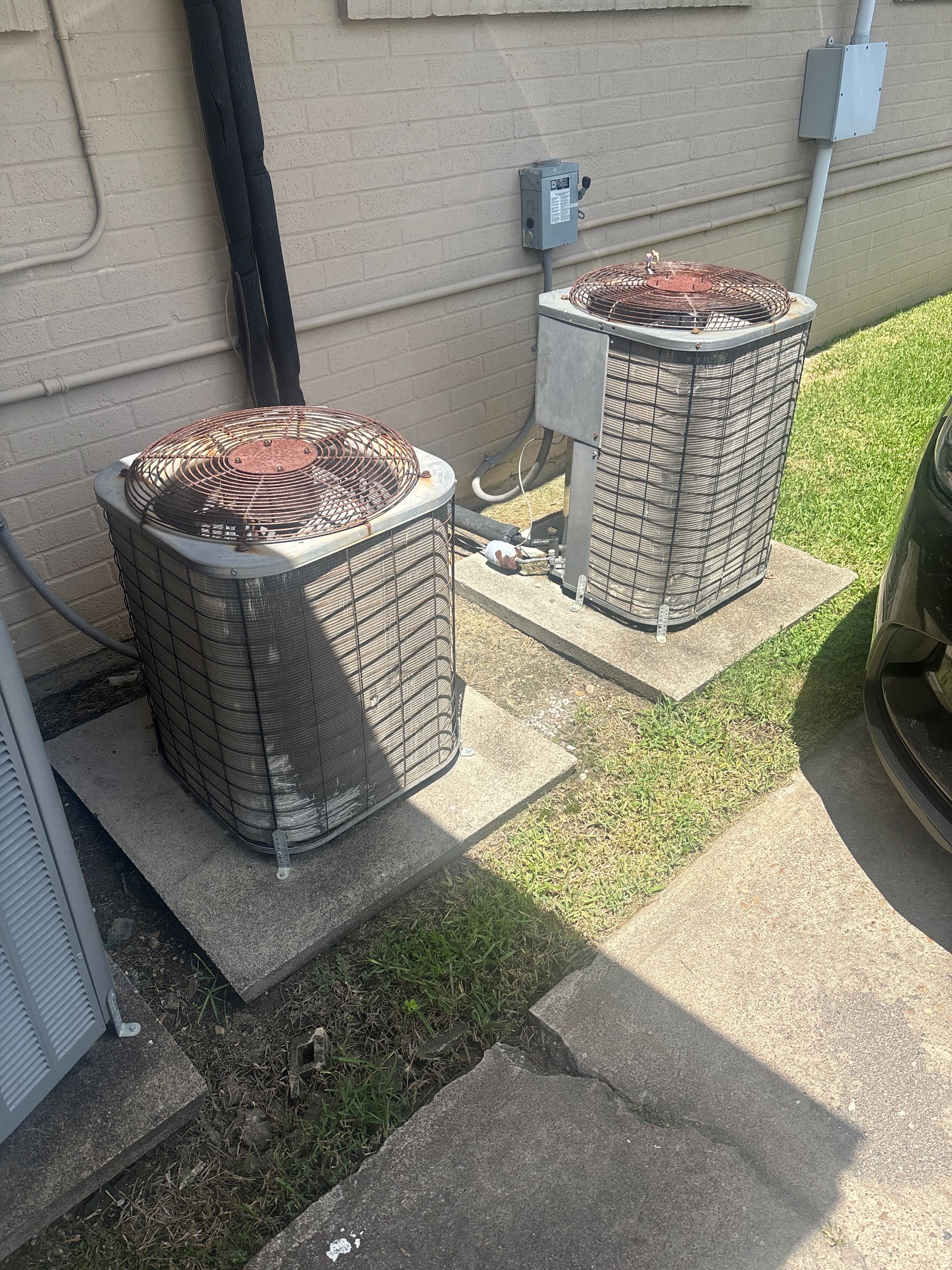 Two outdoor HVAC units beside a building wall on a concrete pad, with grass nearby.