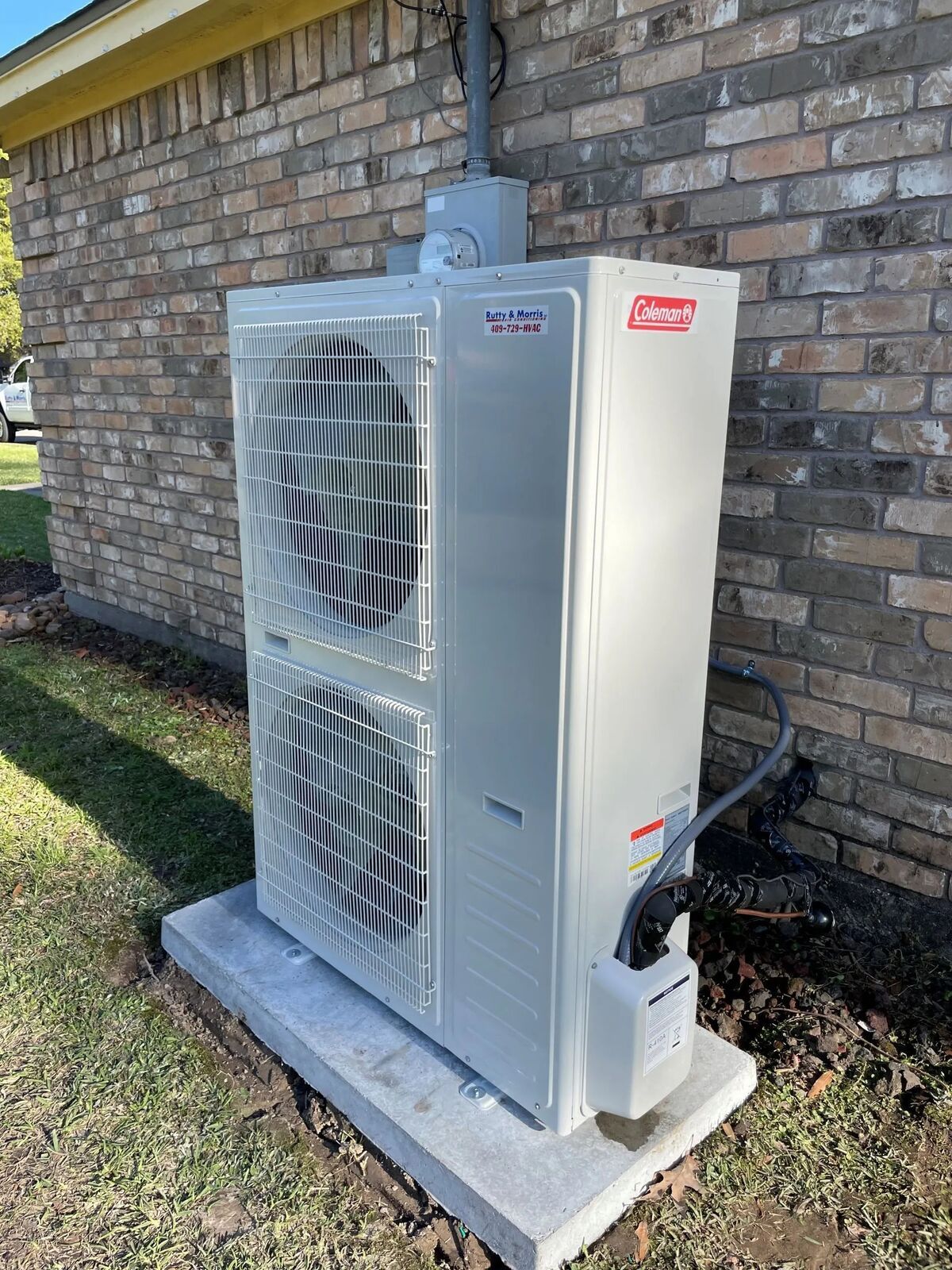 Outdoor HVAC unit beside a brick house wall on a concrete pad, with visible fans and piping.
