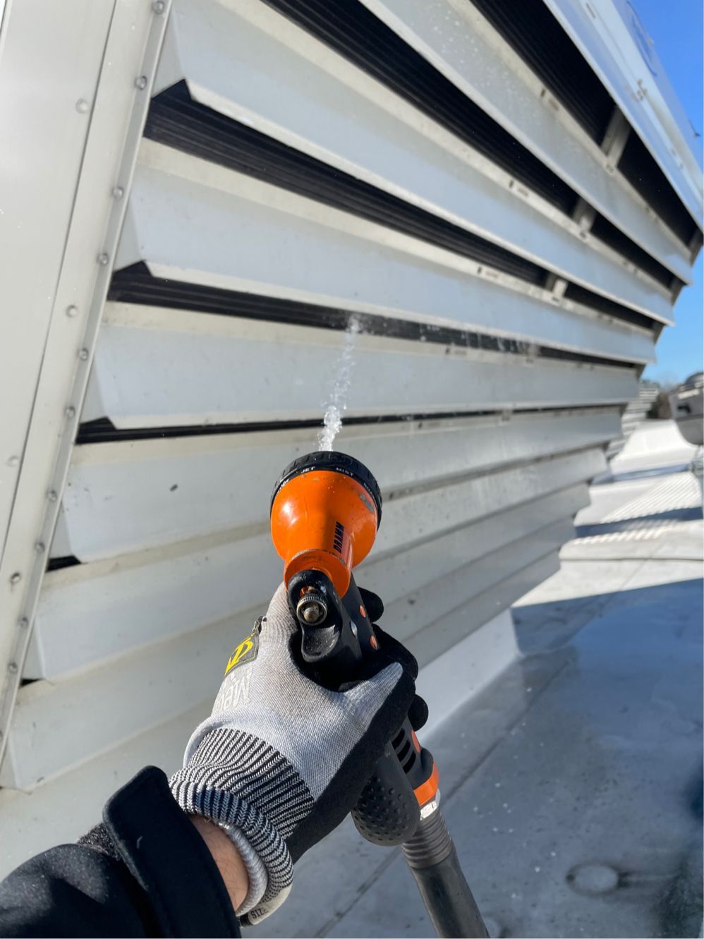 Gloved hand holding an orange spray nozzle aimed at a white vented exterior wall