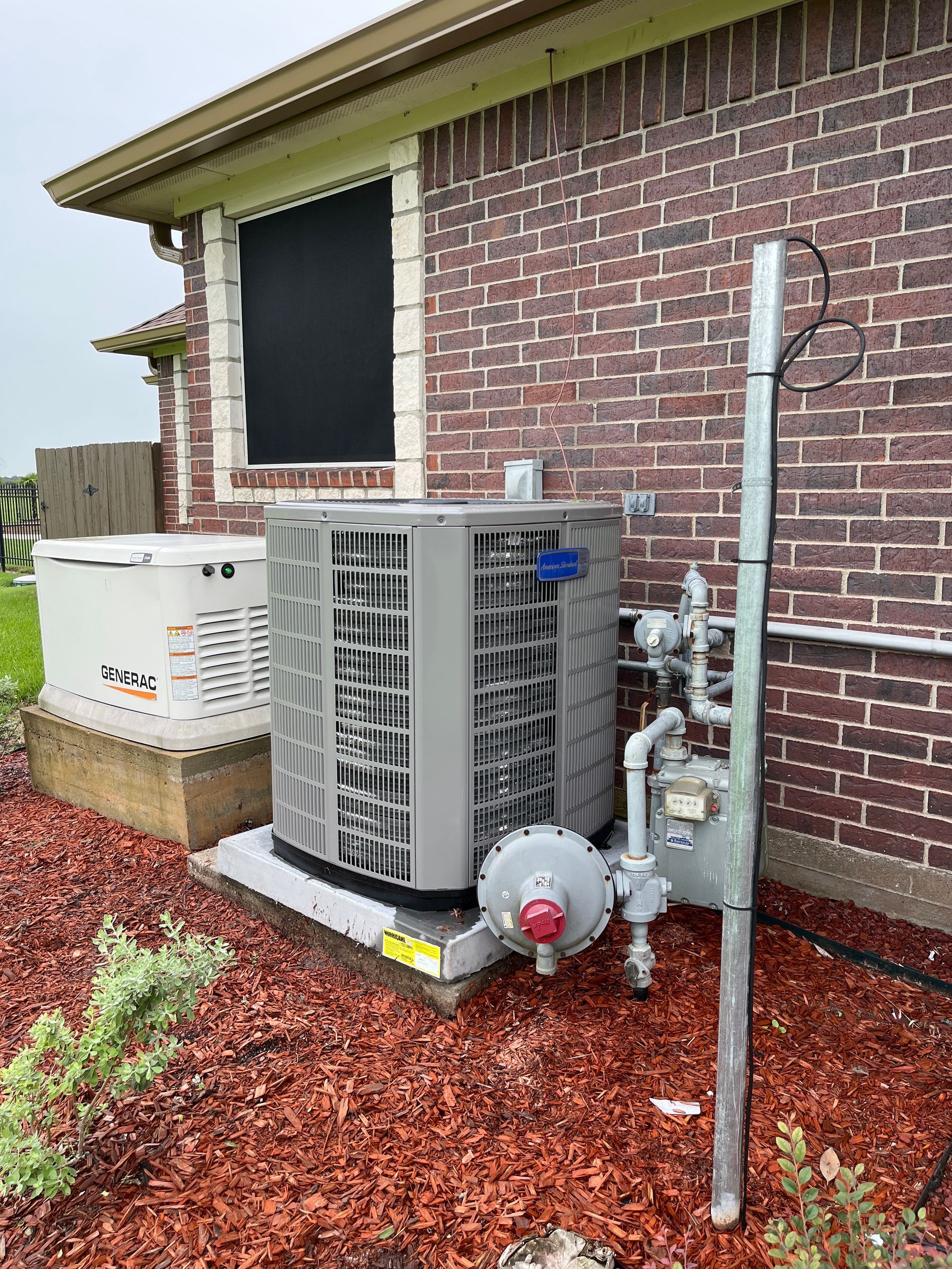 Outdoor HVAC unit beside a brick house, with red mulch and a window nearby.