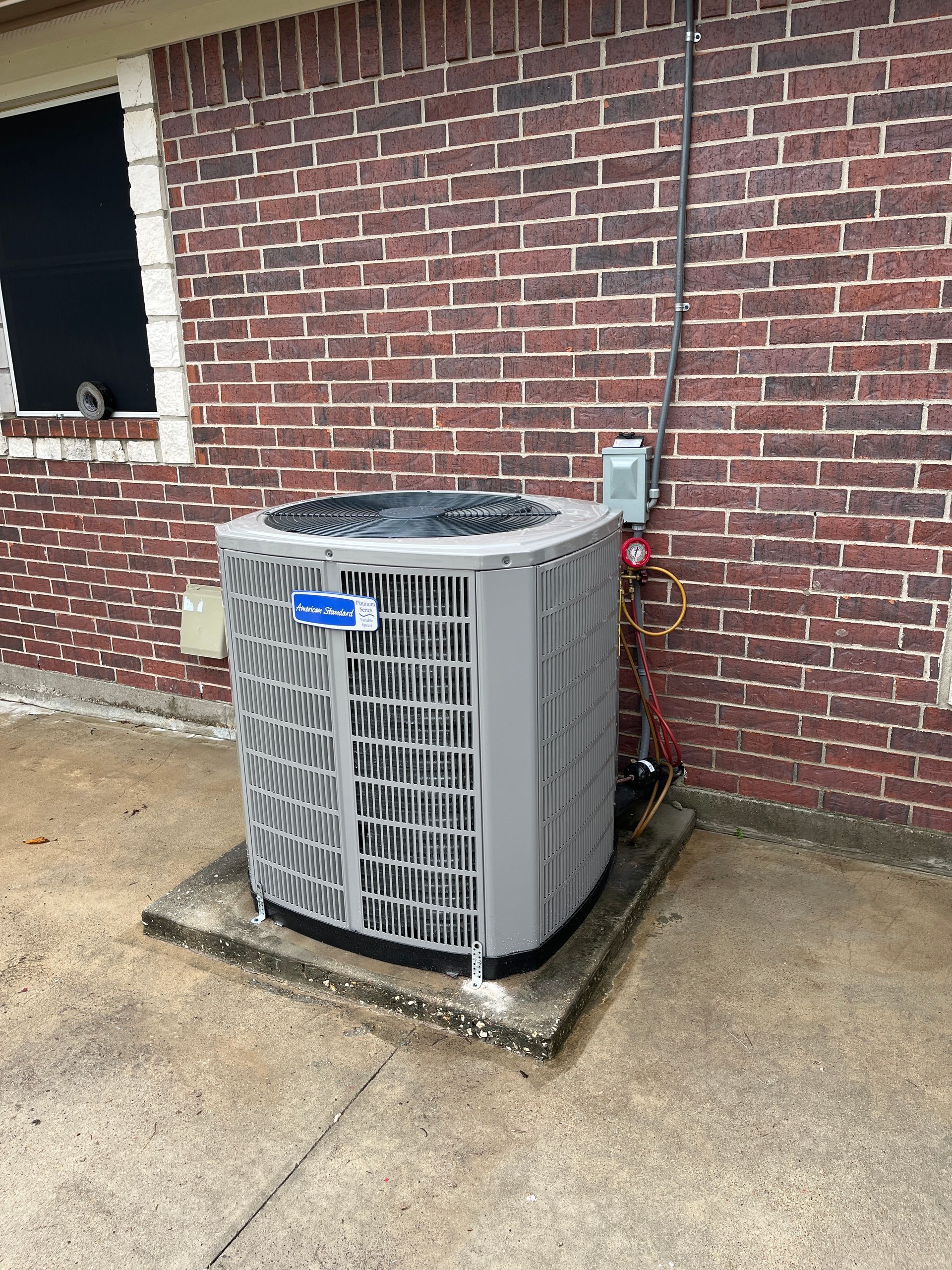 Outdoor air conditioner unit beside a brick wall on a concrete pad, with an open window nearby