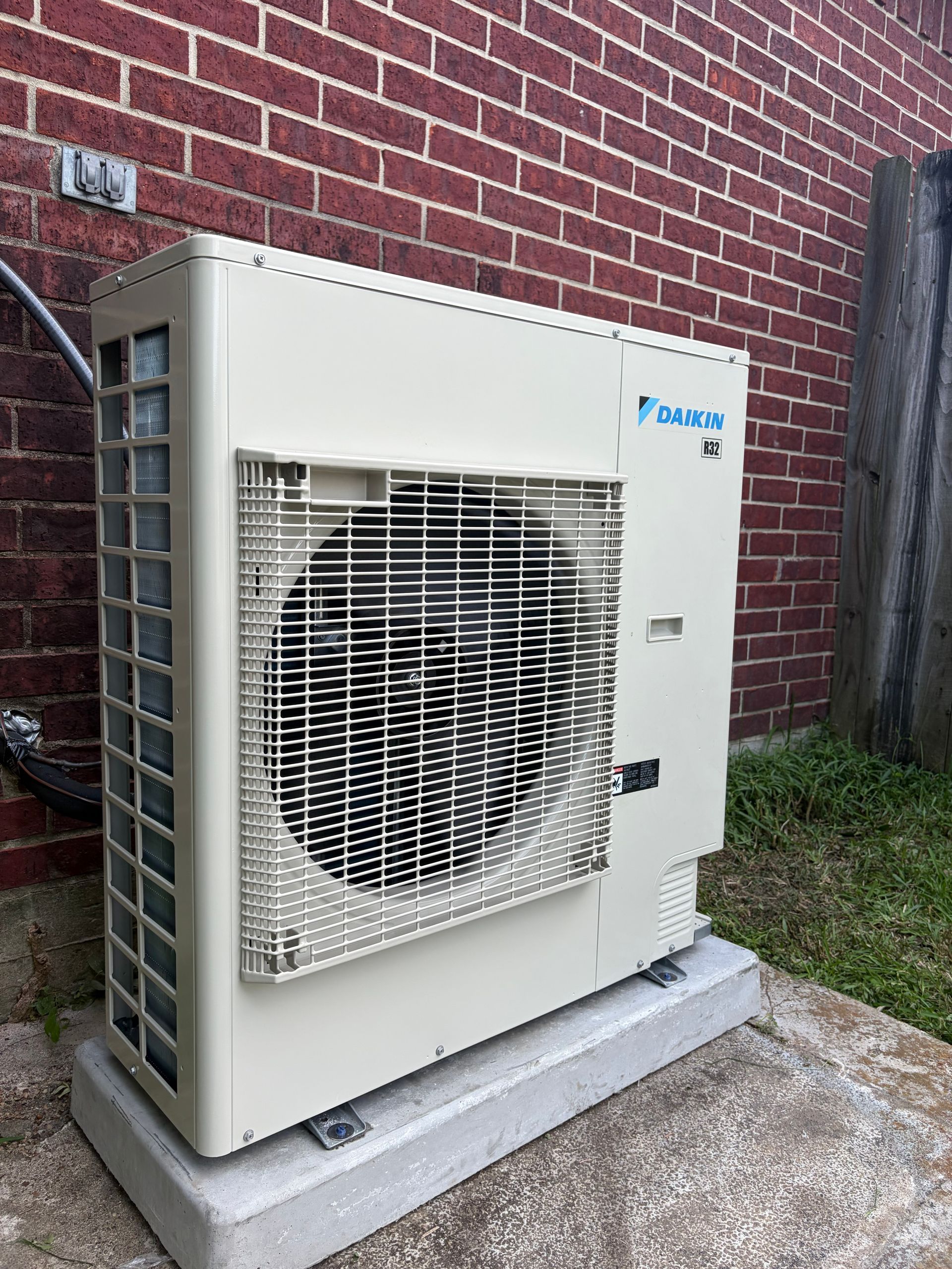 Outdoor Mitsubishi heat pump unit beside a brick wall on a concrete pad, with grass nearby.