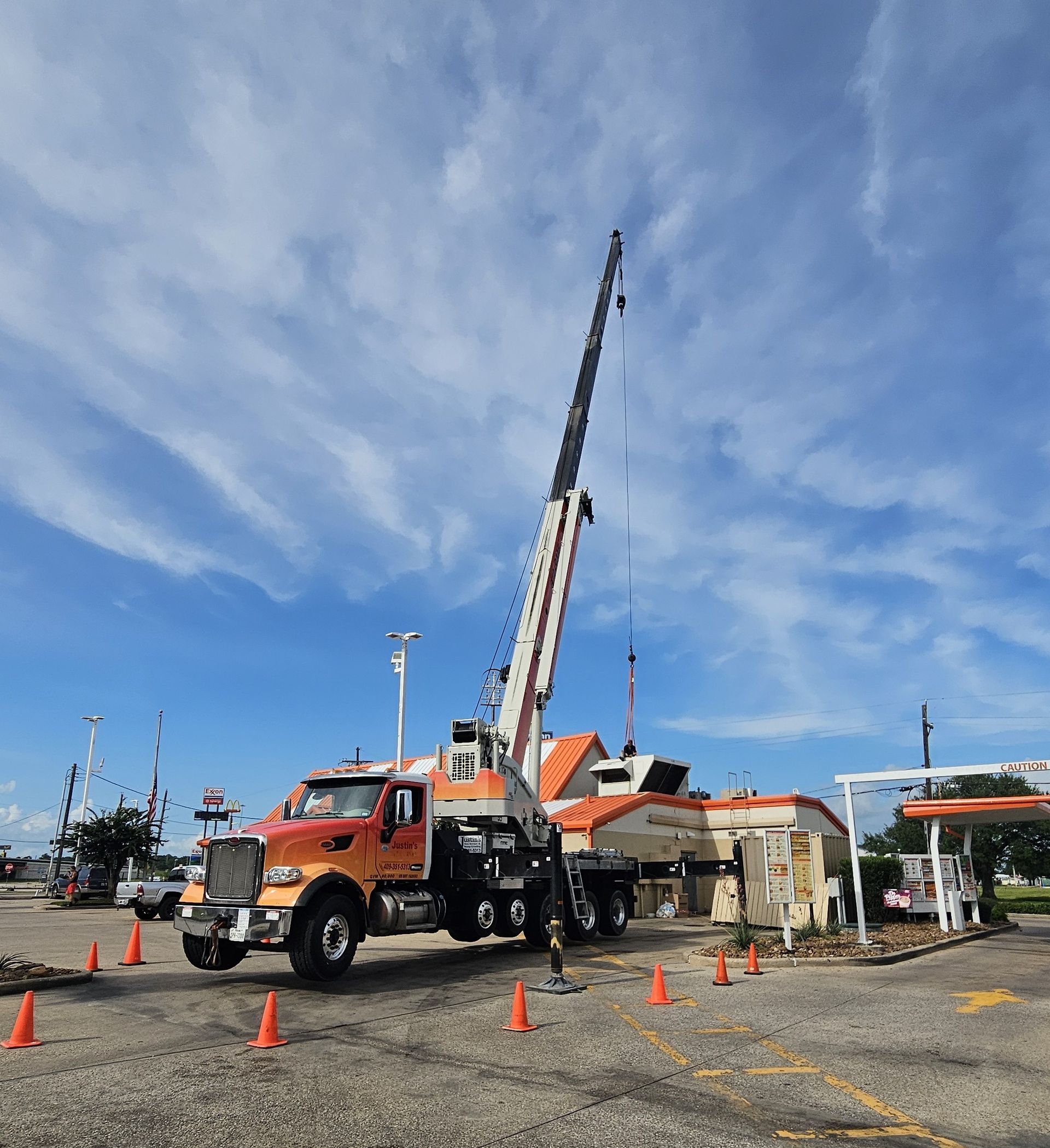 Orange crane truck with extended boom at a construction site under a blue sky, surrounded by cones