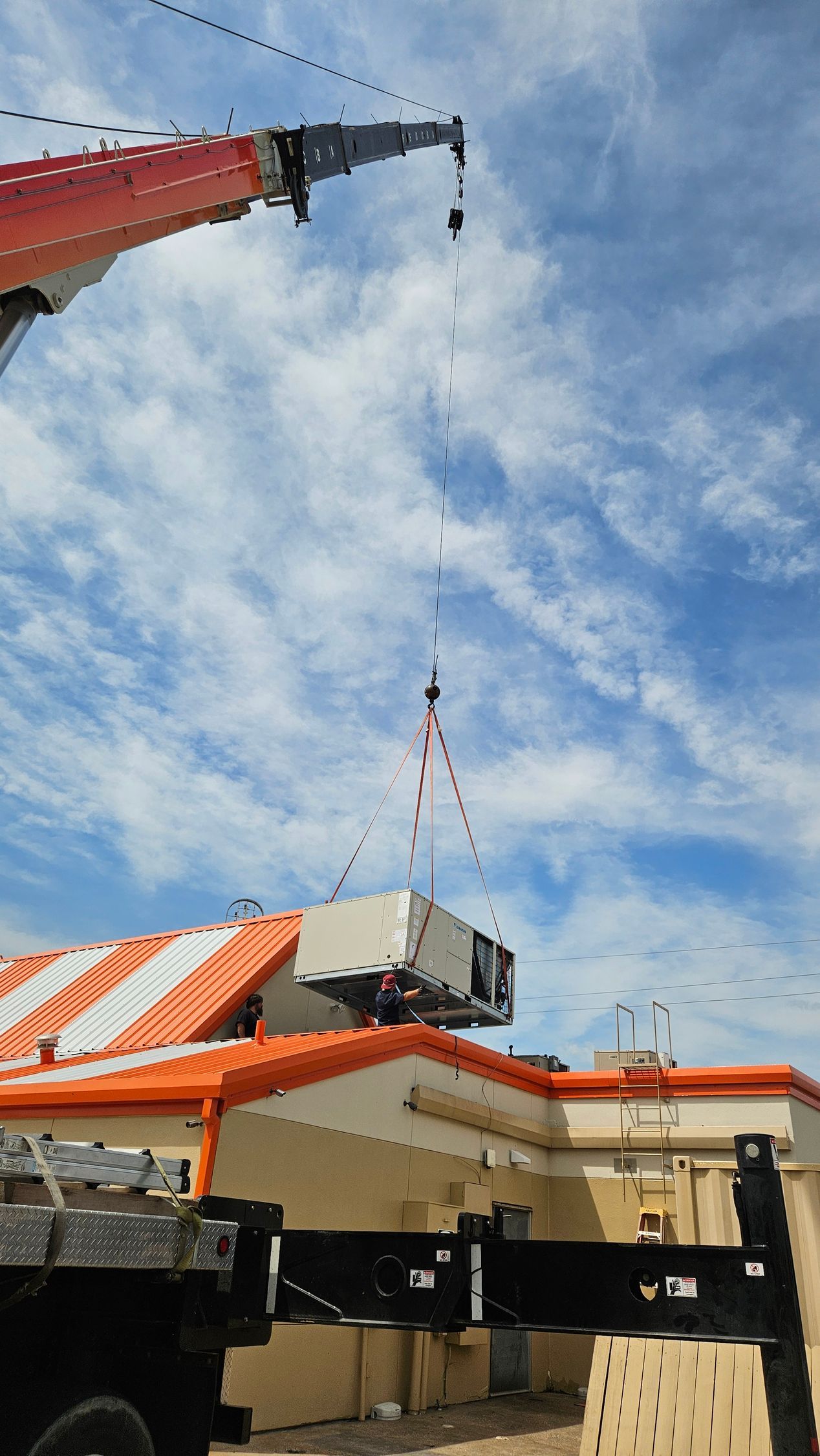 Crane lifting a white rooftop unit onto an orange-roofed building under a blue sky.