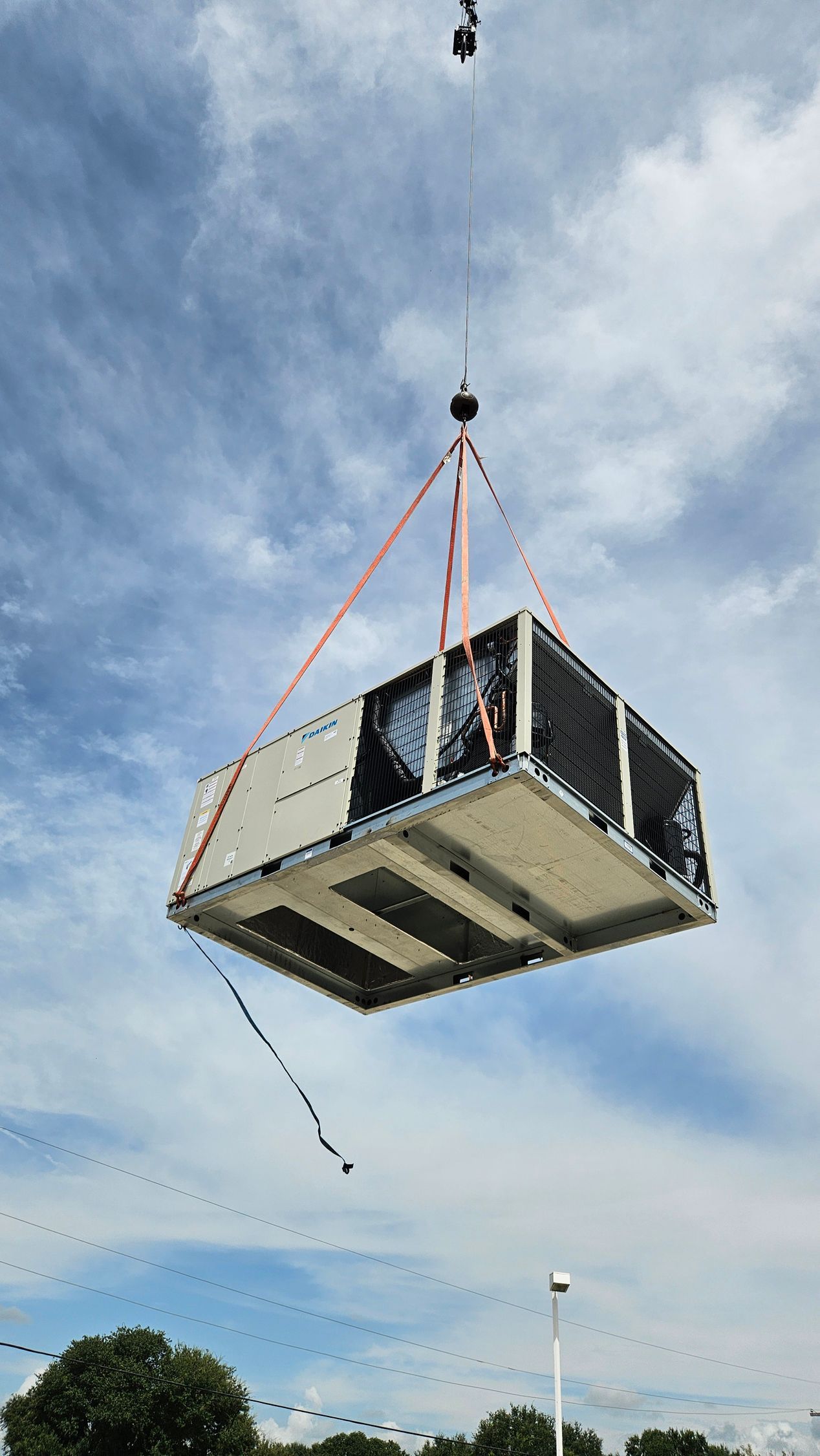 Large metal box suspended by crane cables against a cloudy sky above trees and a streetlight