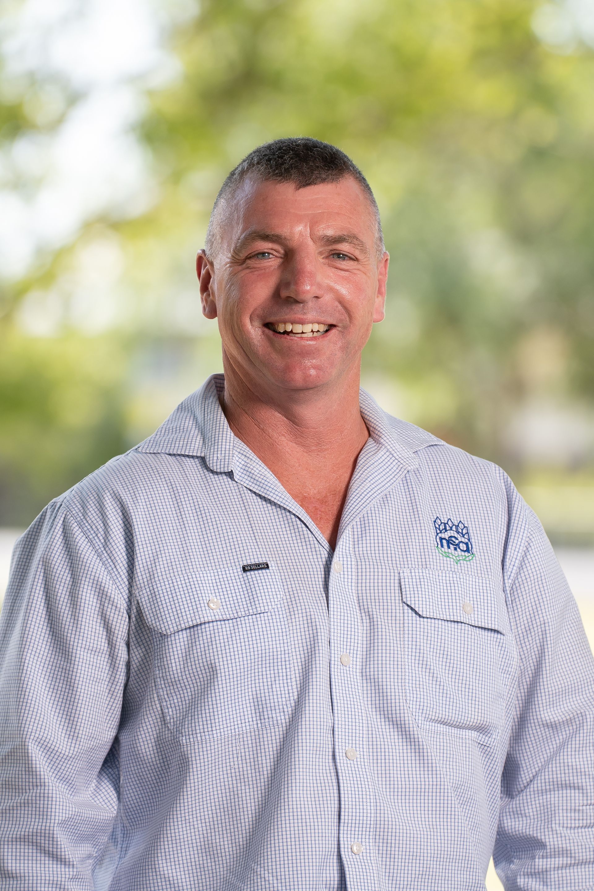 Man wearing a blue collared shirt smiles outdoors, with a blurred background of trees and light.