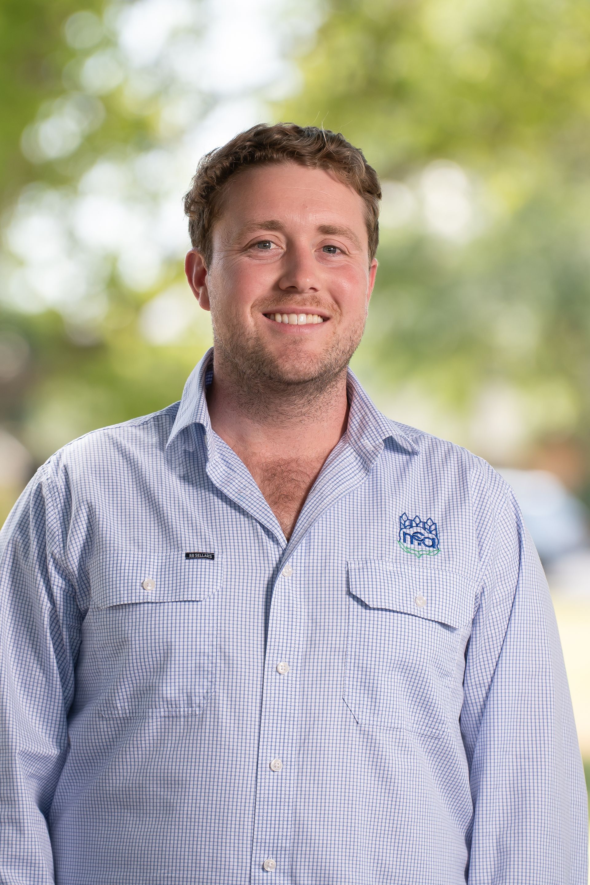 Man wearing a blue and white striped shirt smiles outdoors, with a blurred green background.